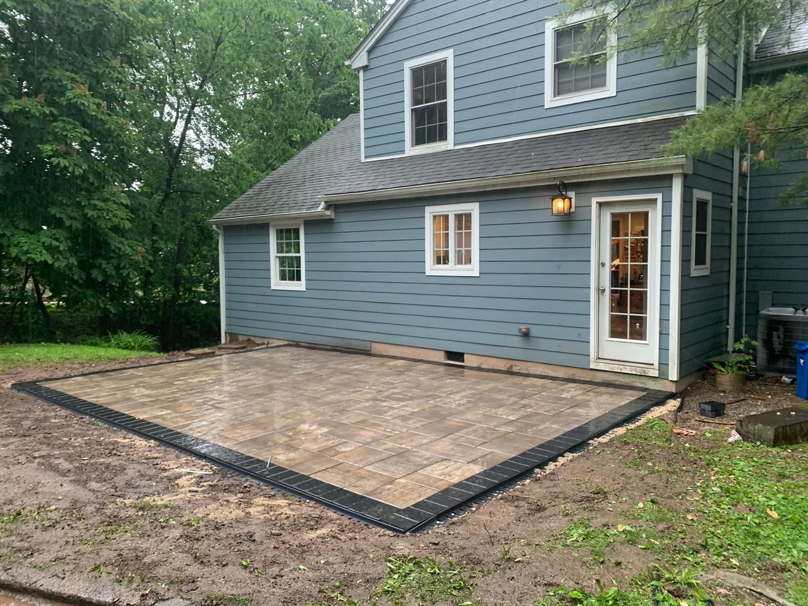 Backyard patio with gray pavers and dark border next to a blue house with white trim and a door.