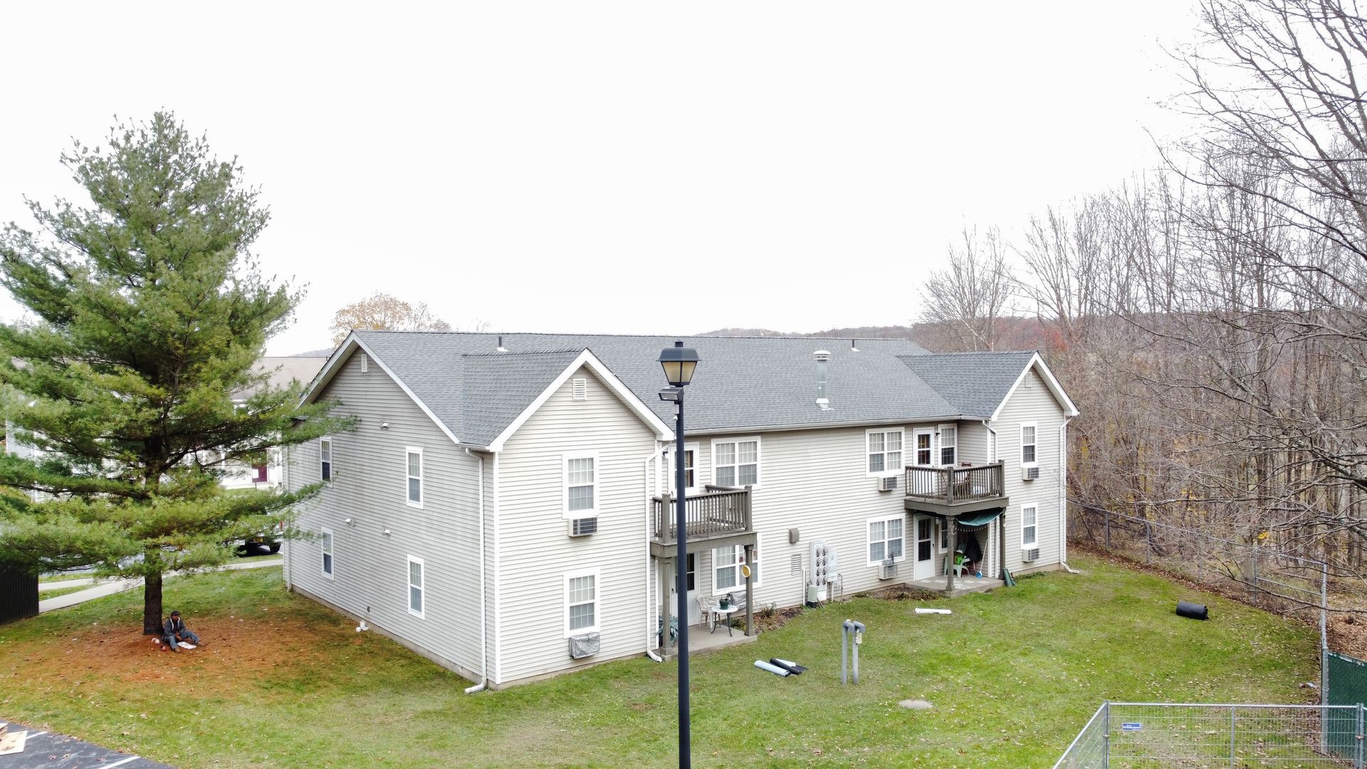 Two-story light-gray apartment building with balconies, surrounded by green grass and trees. Overcast day.