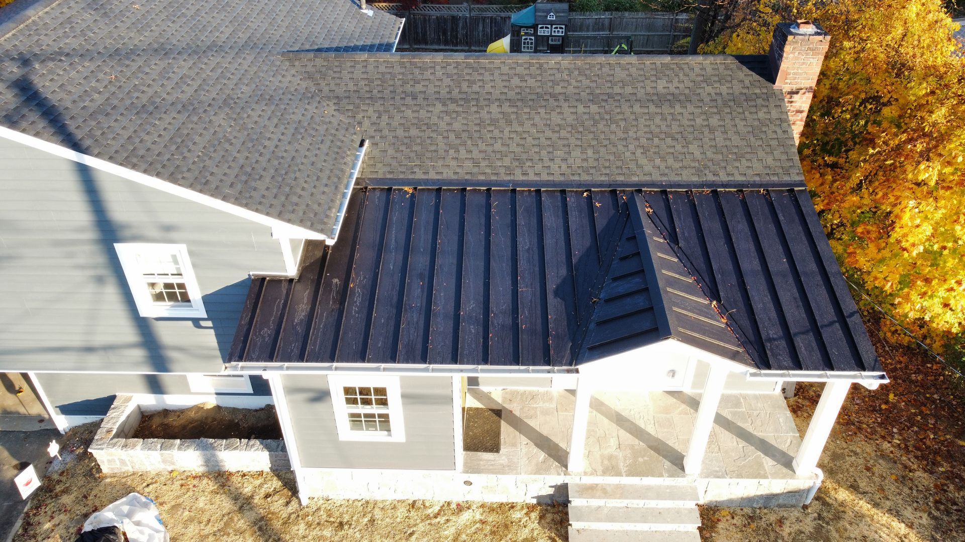 Overhead view of a gray house with a black metal roof, autumn trees in background.