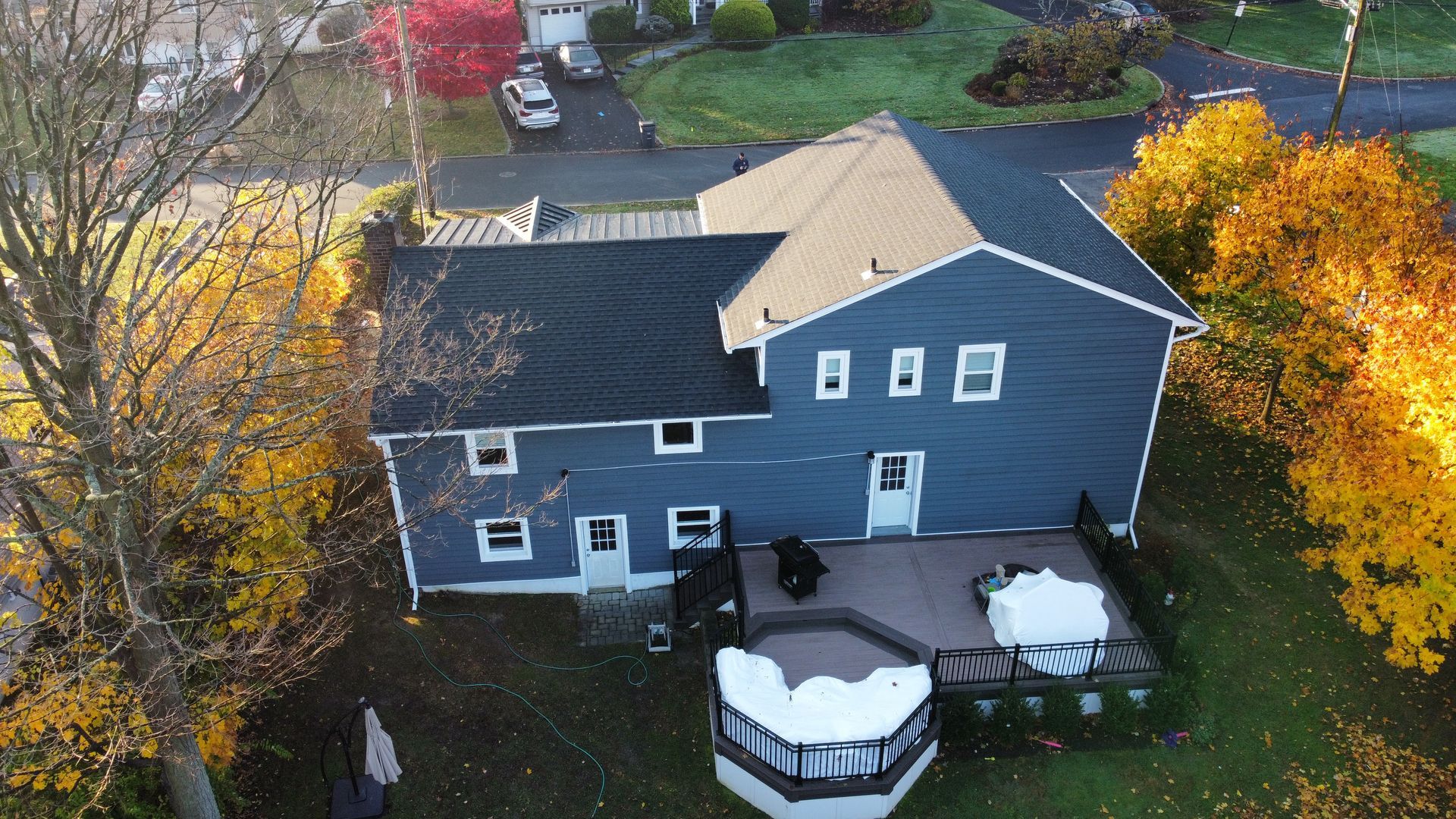 Two-story blue house with deck, surrounded by fall foliage, cars in driveway.