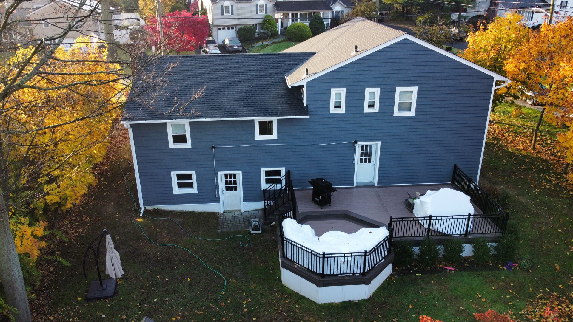 Blue-sided two-story house with a deck and hot tub in a yard surrounded by fall foliage.