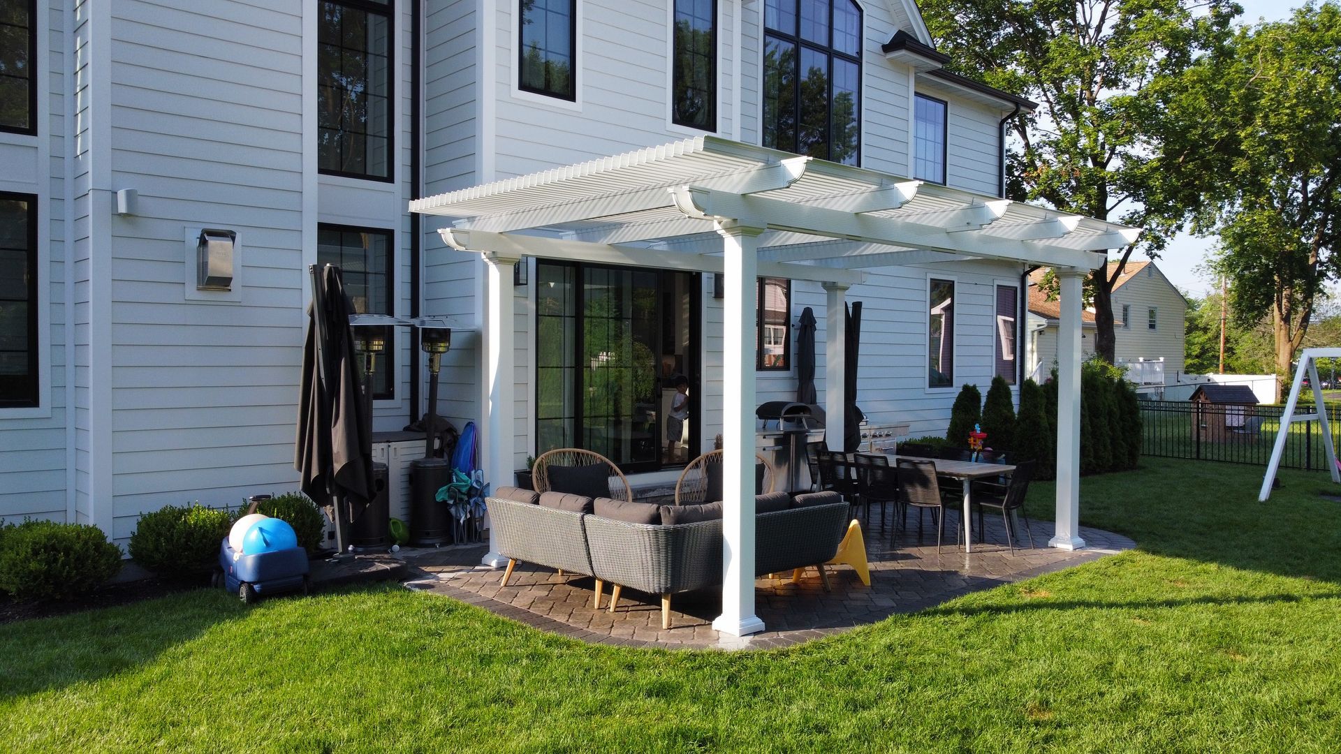 White pergola over a patio with outdoor furniture next to a white house with black windows, on a grassy lawn.