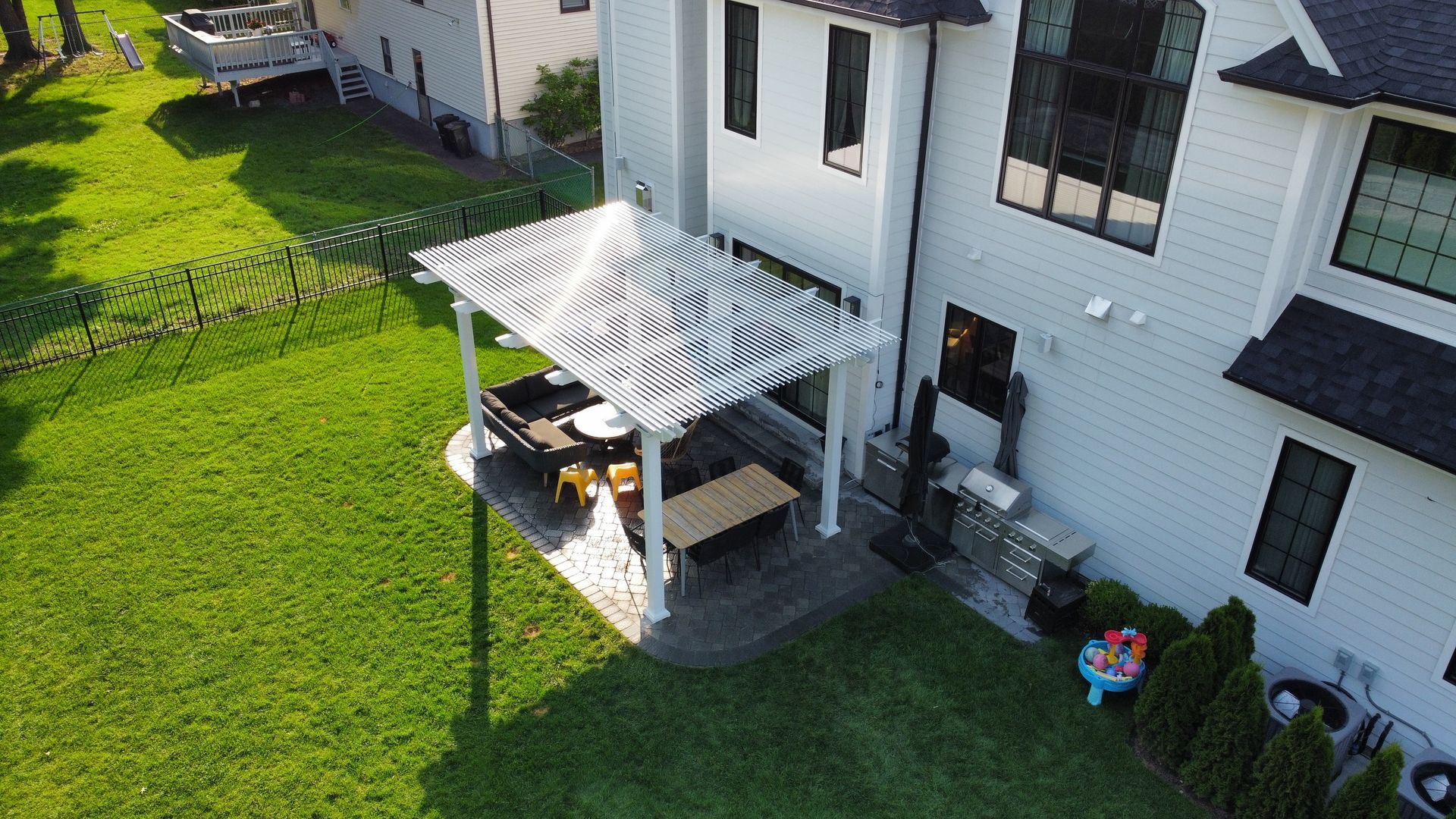 Backyard with a gray-sided house, patio, pergola, table, and chairs. Lush green grass and blue sky.
