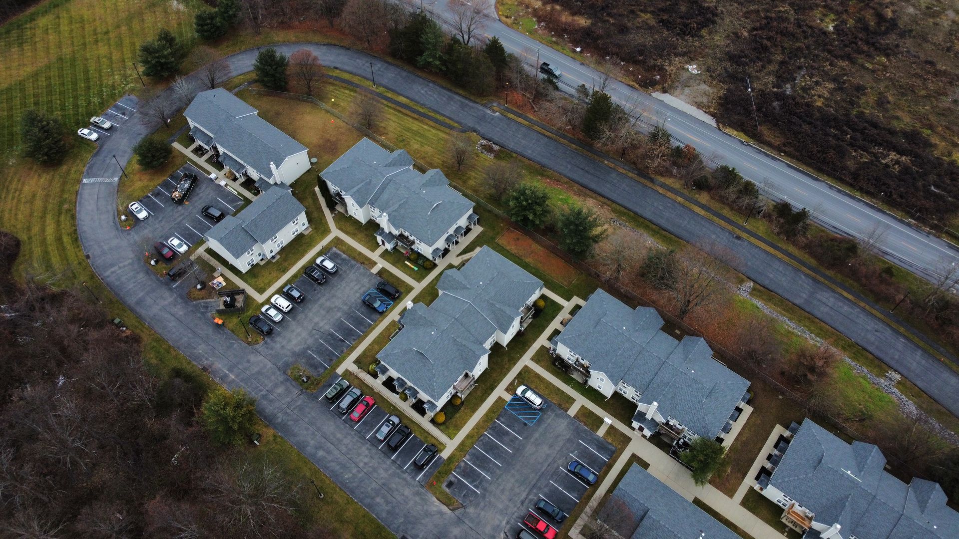 Aerial view of an apartment complex with parking, surrounded by trees and a winding road.