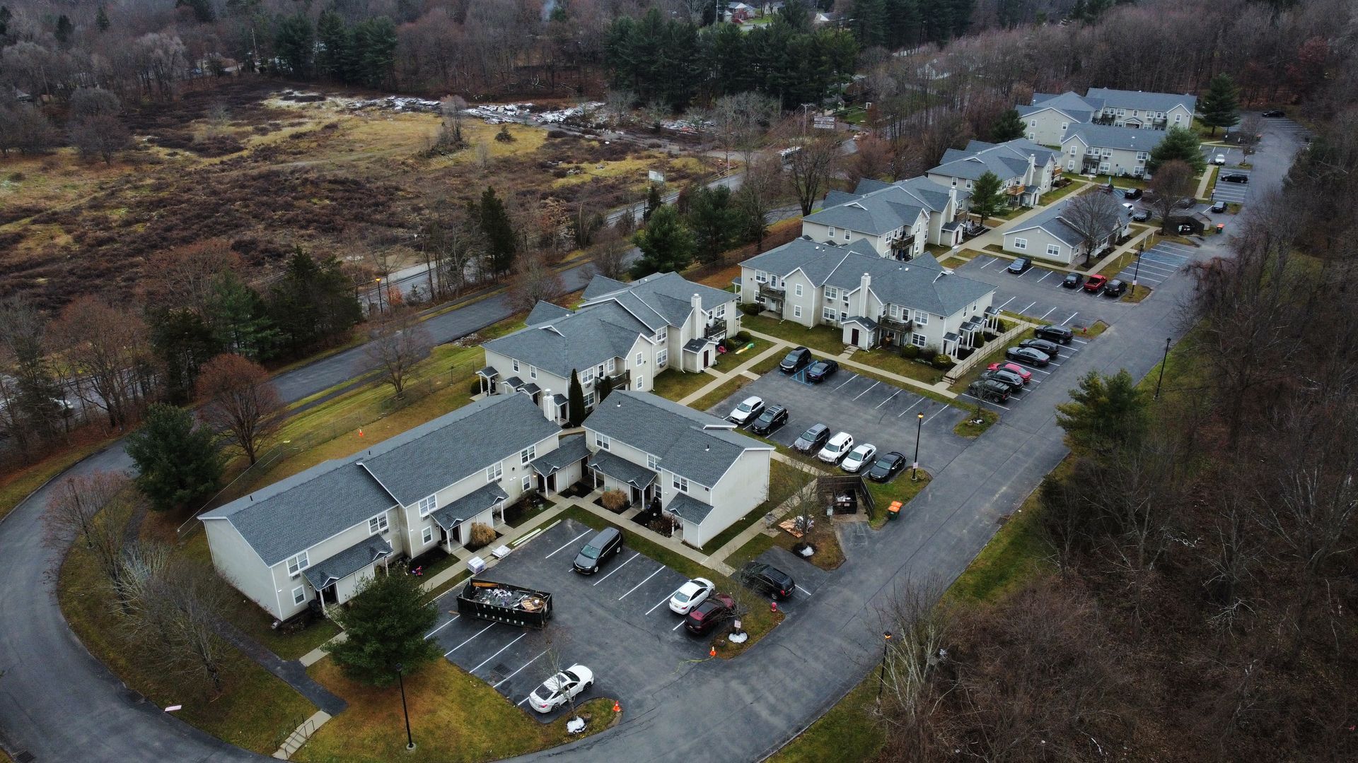 Aerial view of gray apartment buildings with parking in a wooded area.