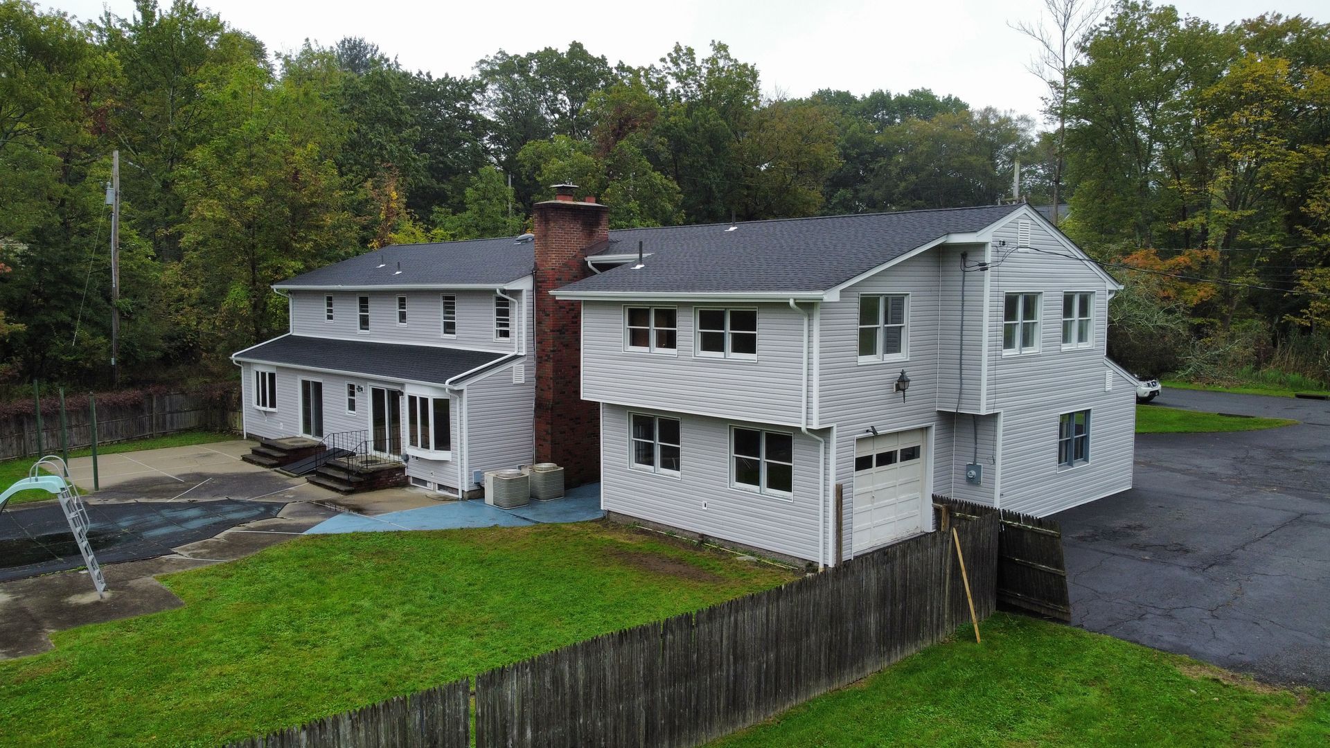 Two-story gray house with white trim, brick chimney, and a garage. Green lawn and trees surround it.