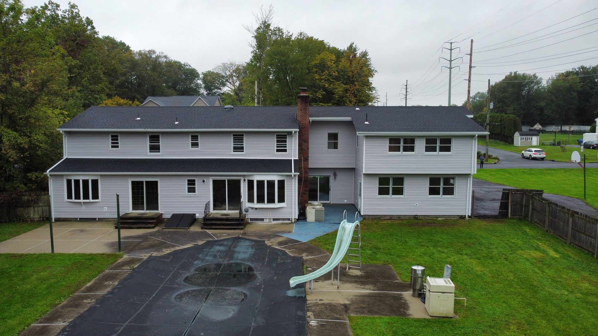 Backyard of a two-story house with a dark roof, pool, and green yard; cloudy sky.