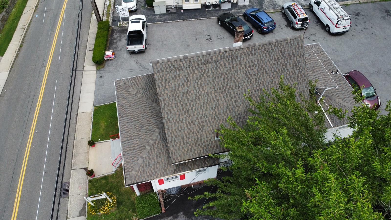 Aerial view of a building with a gray roof next to a parking lot with cars, near a road.