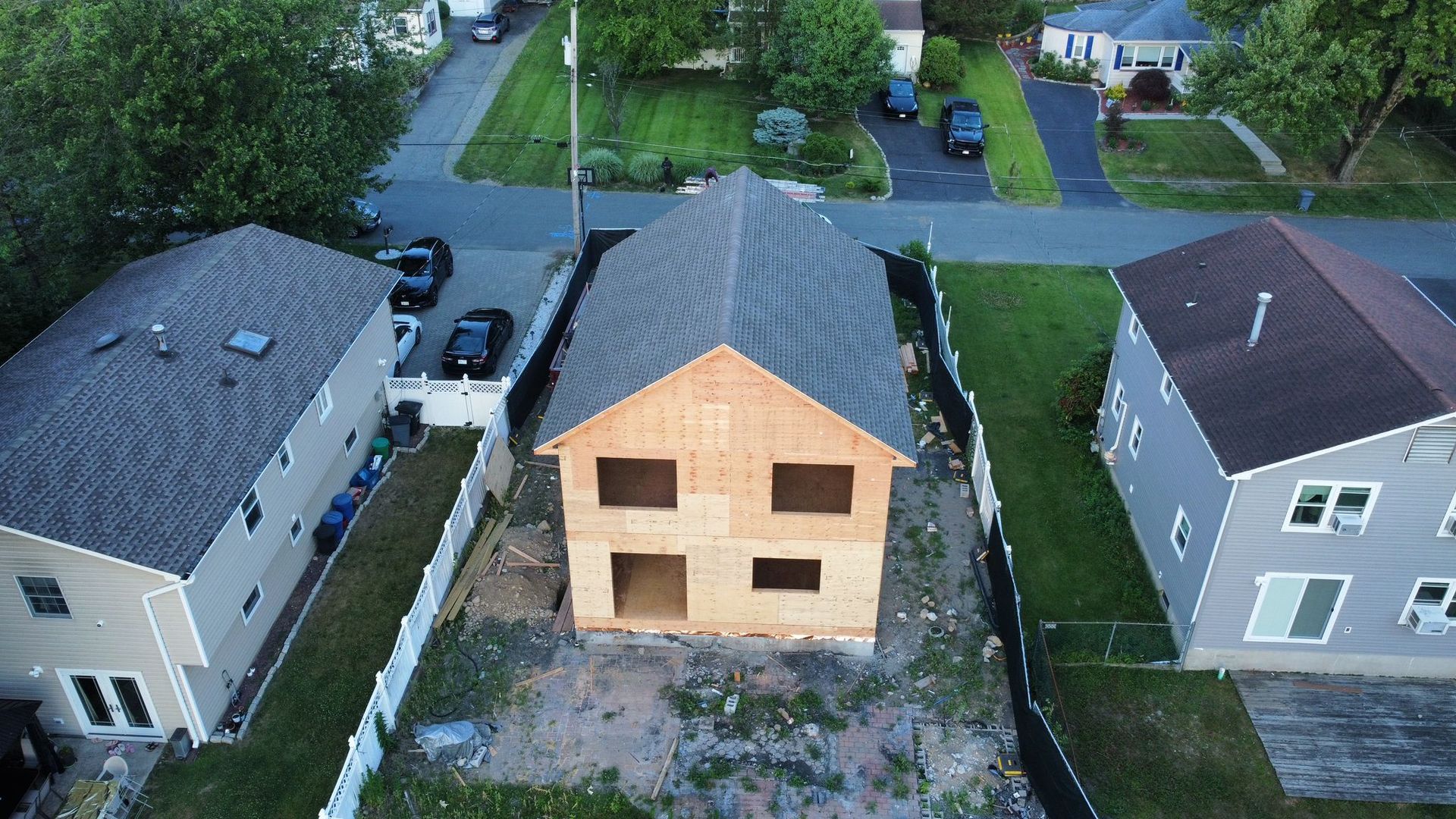 Two-story house under construction between two finished houses, green lawns, gray asphalt, and a blue sky.