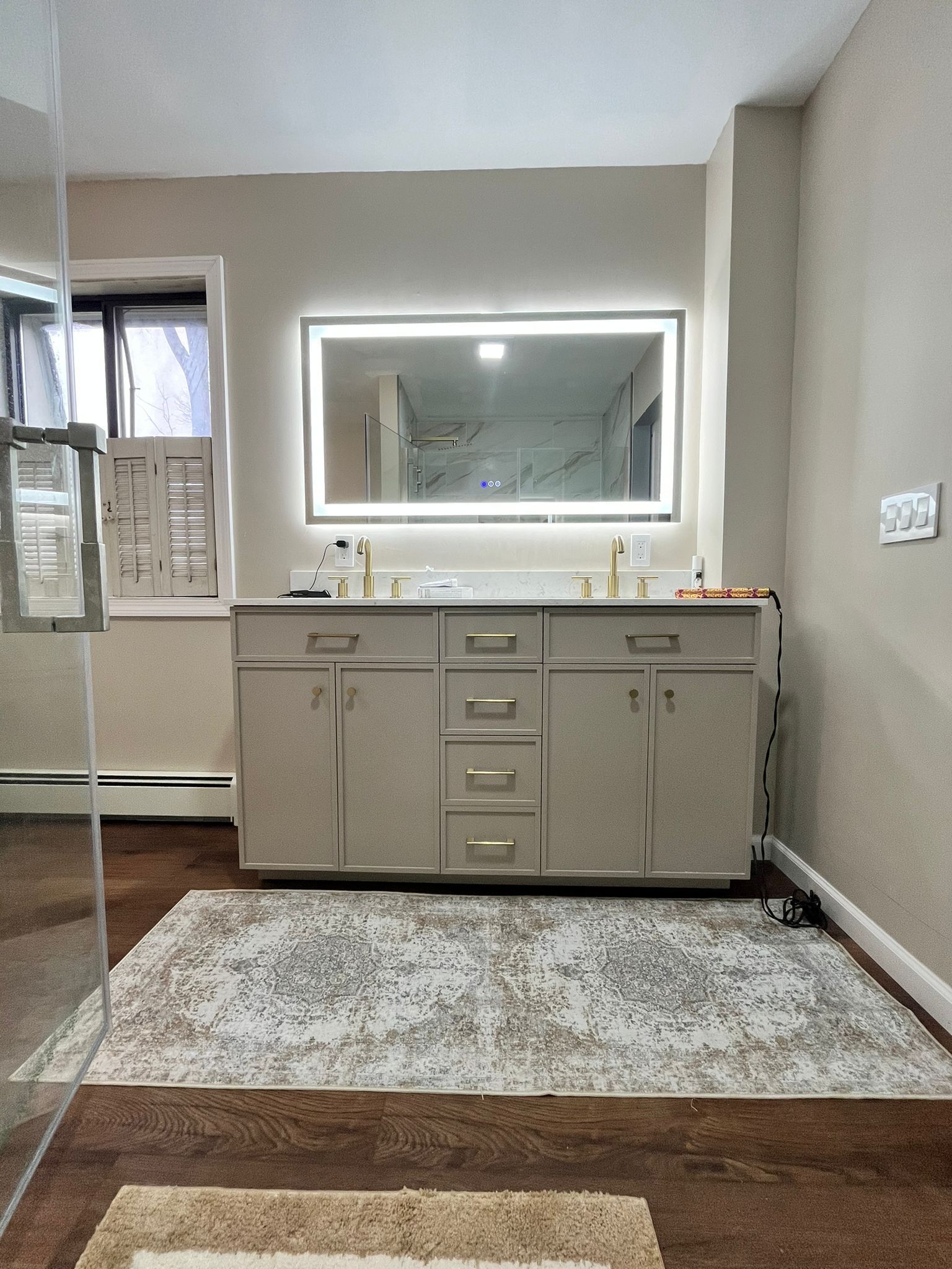 Bathroom with beige vanity, lighted mirror, window, and patterned rug on wooden floor.