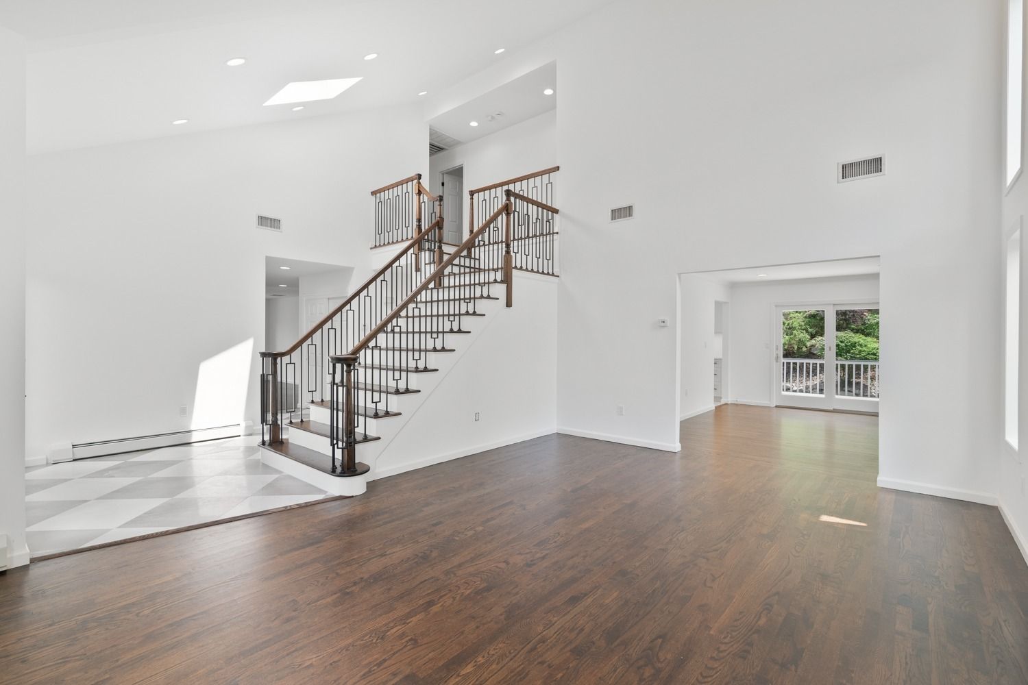 Empty, white-walled room with dark wood floors and staircase. Natural light from windows and skylight.