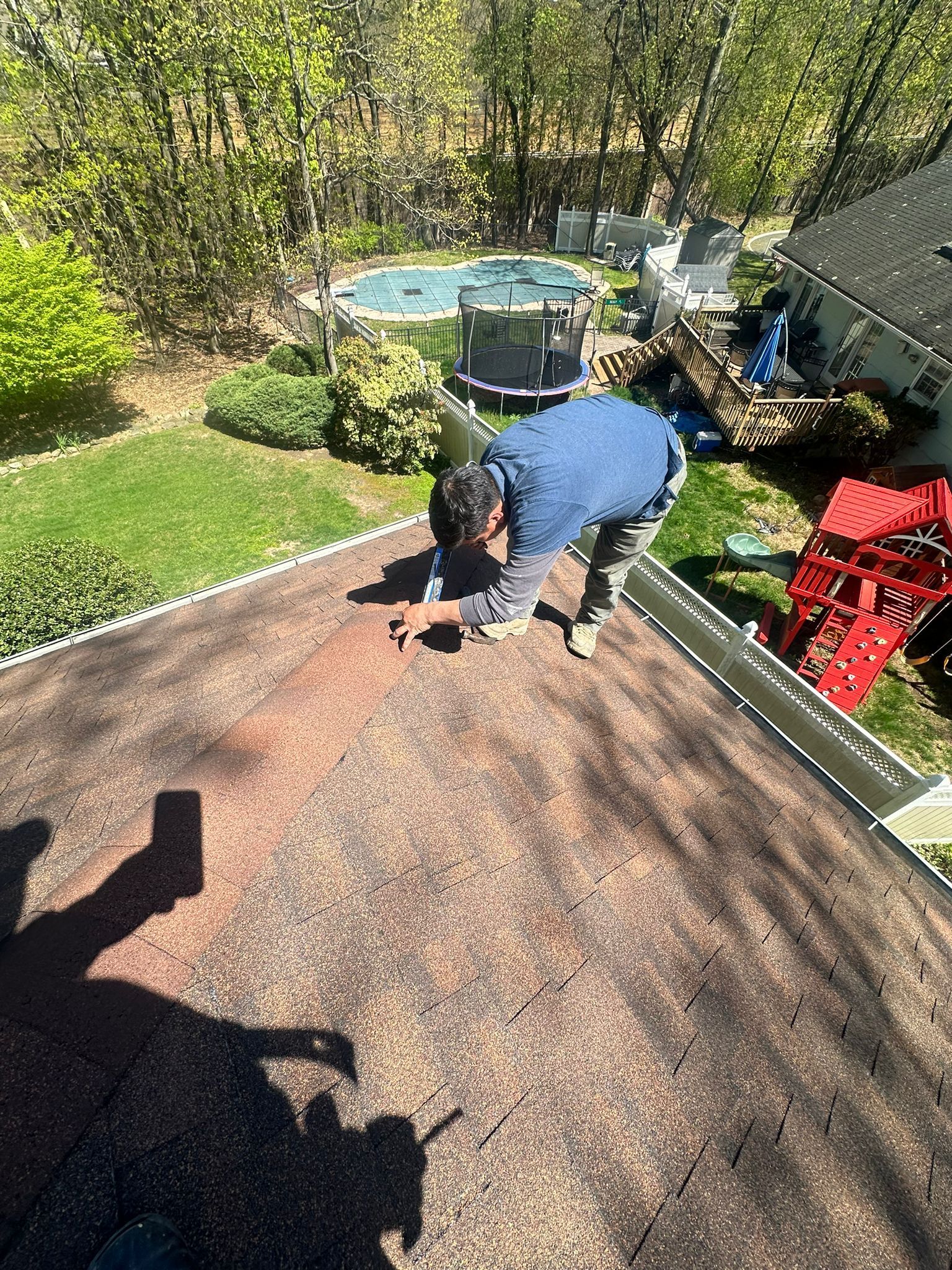 Person on a roof repairs it. Brown shingles, green yard, a pool, blue shirt. Bright sunny day.