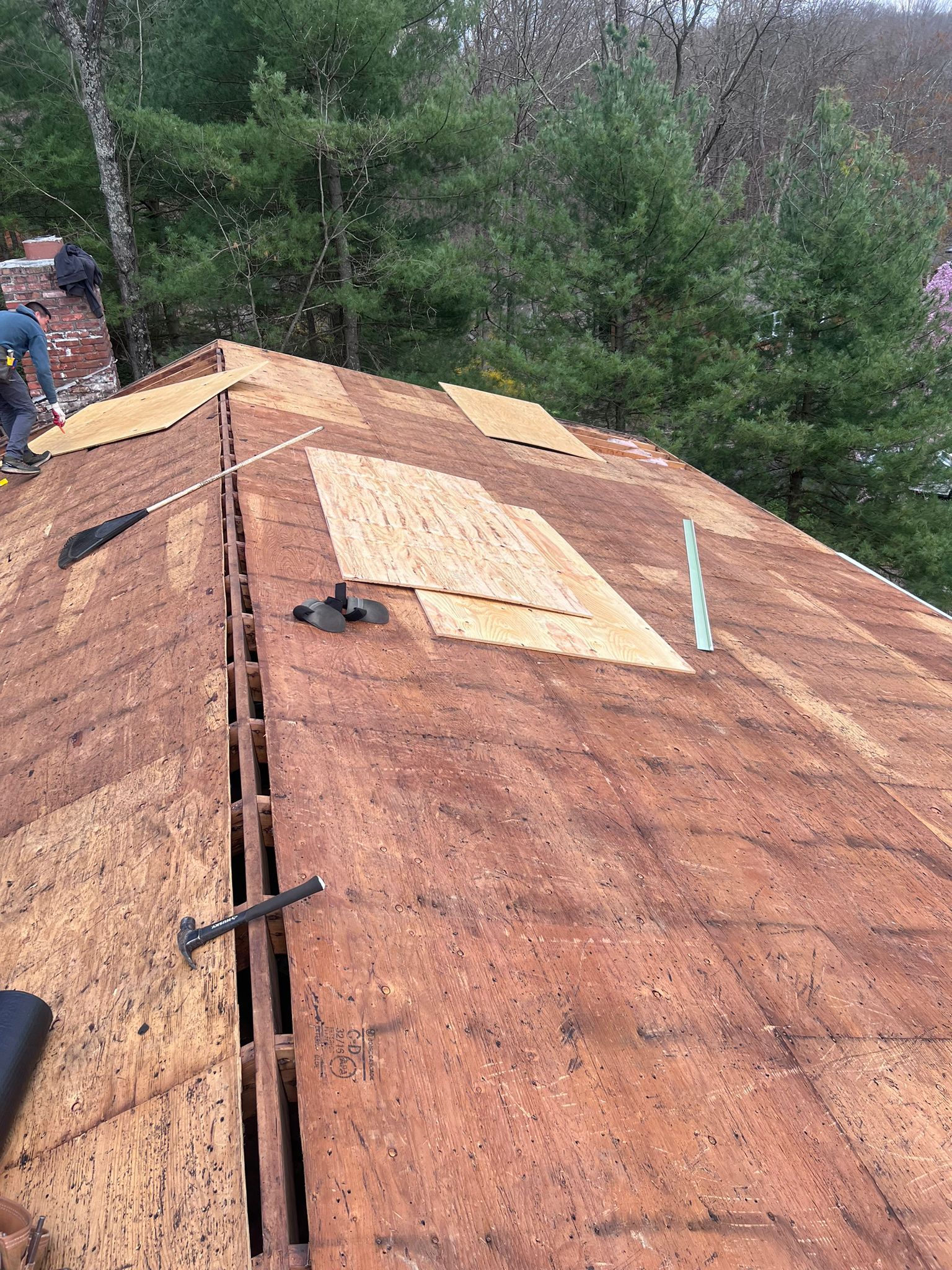 Roof partially stripped of shingles, revealing wood. Roofer works near a brick chimney and trees.