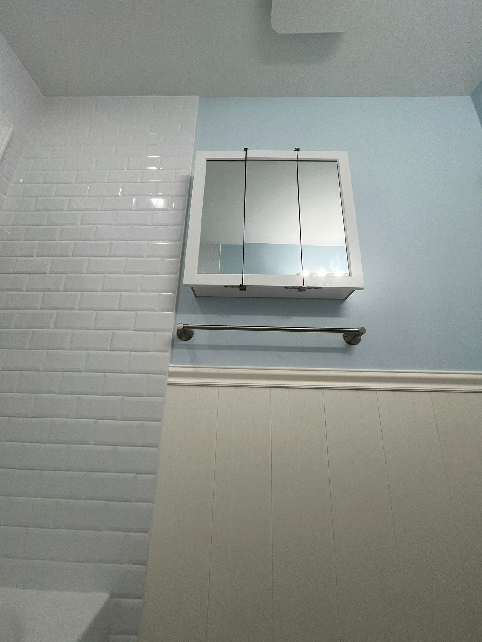 White tiled wall to the left, a medicine cabinet, towel rack on blue wall, and off-white wainscoting.