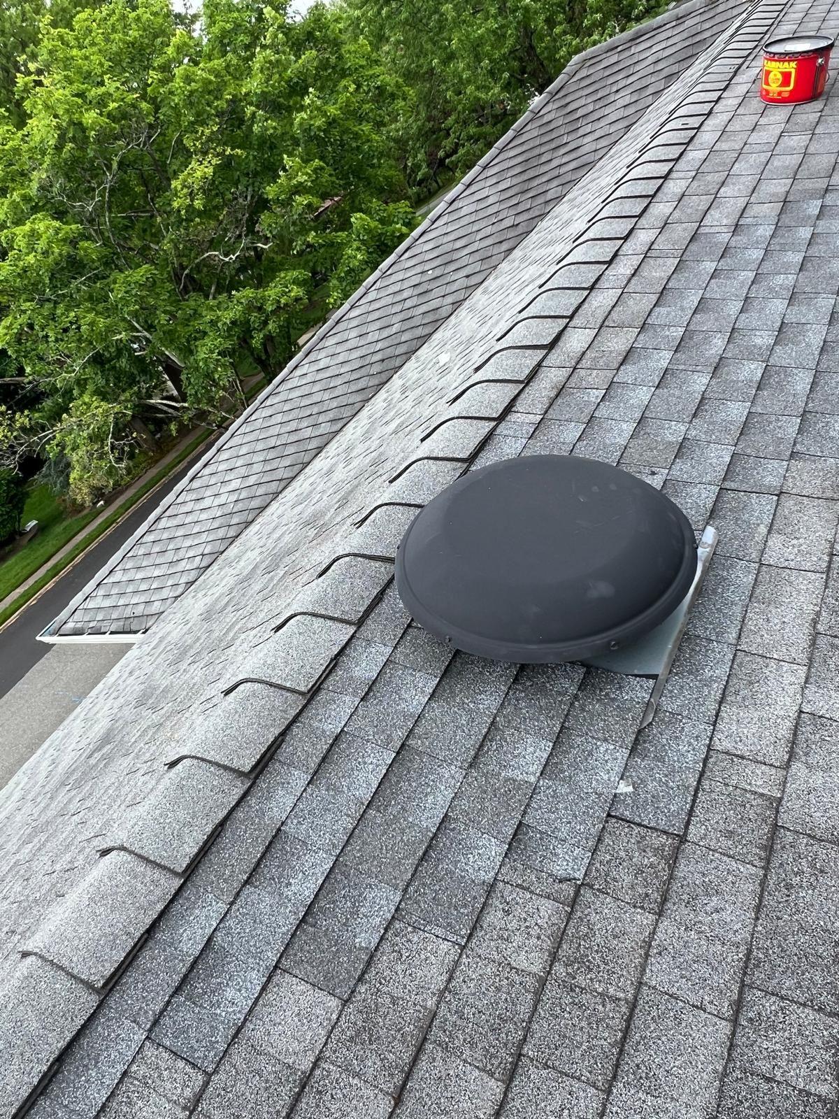 Gray shingled roof with a dark, circular vent and red object in the distance against a backdrop of trees.
