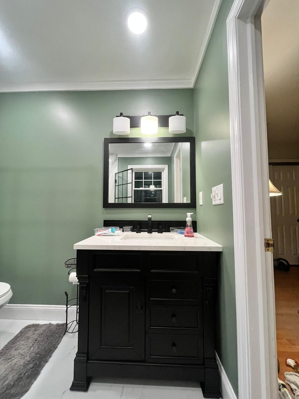 Black vanity with white countertop, rectangular mirror, and three-light fixture on a green wall in a bathroom.