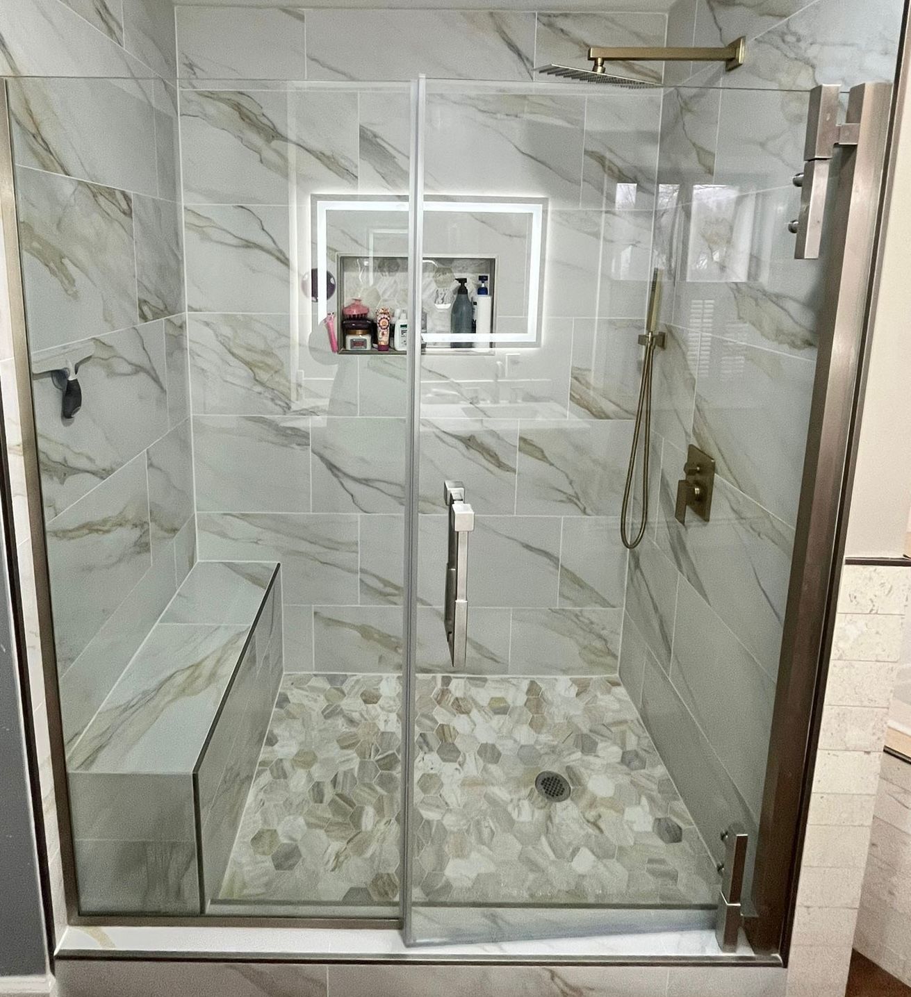 Modern shower with marble-patterned tiles, glass doors, built-in bench, and pebble floor.