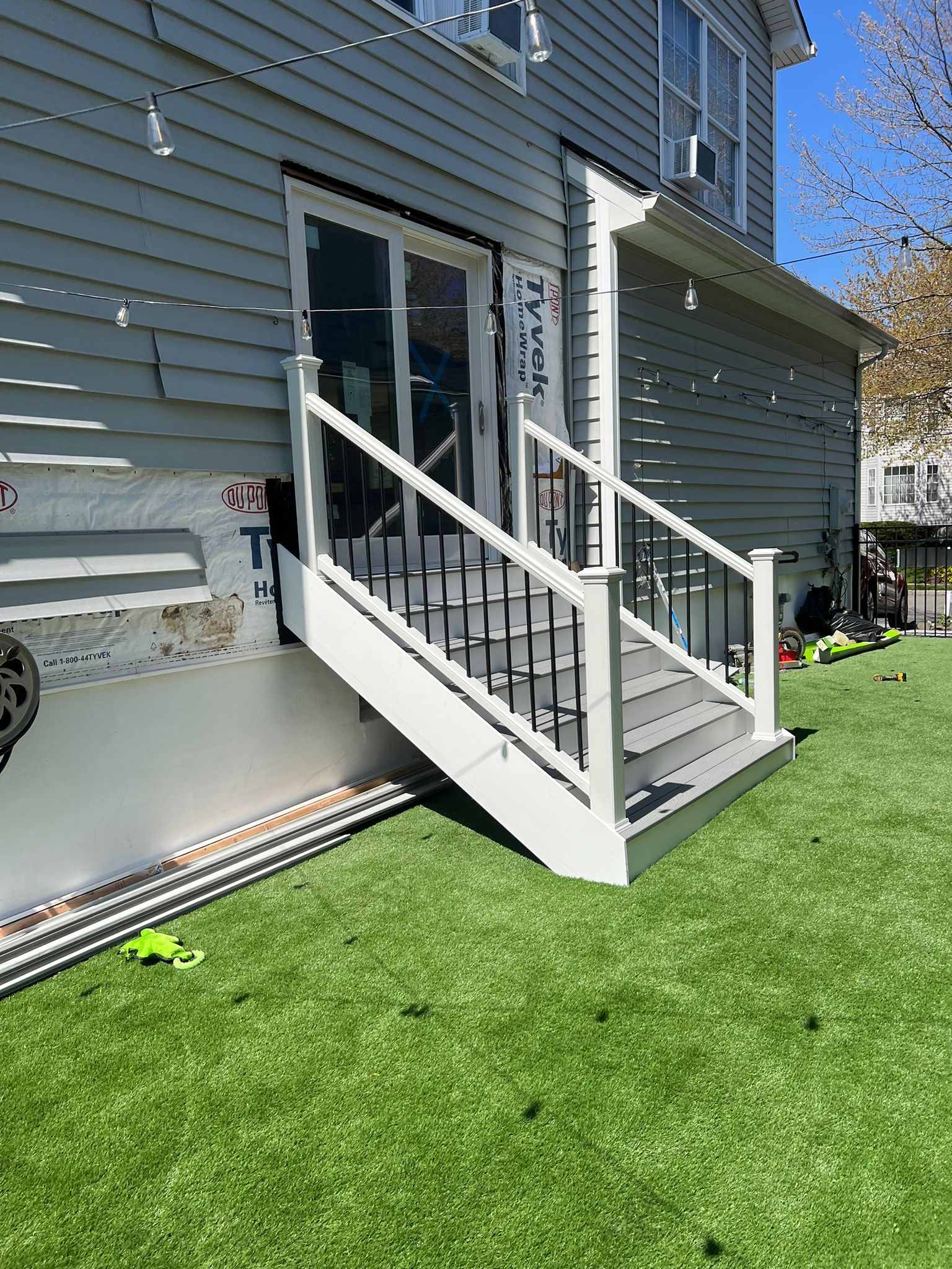 Gray house with white deck and black railing. Steps lead to a glass door, on green grass.