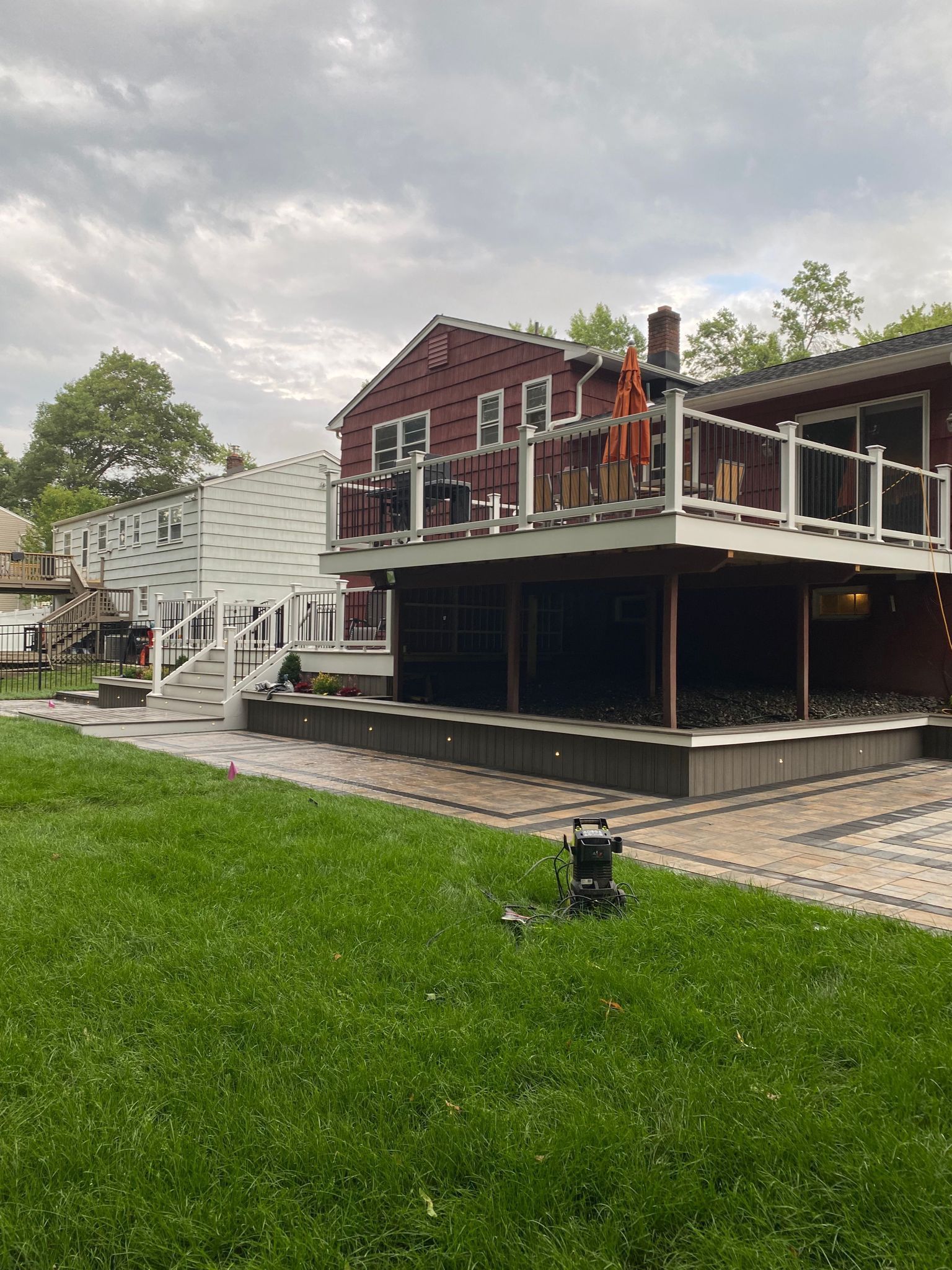 Red house with multi-level deck, glass railings, stairs, and brick patio, under cloudy sky.