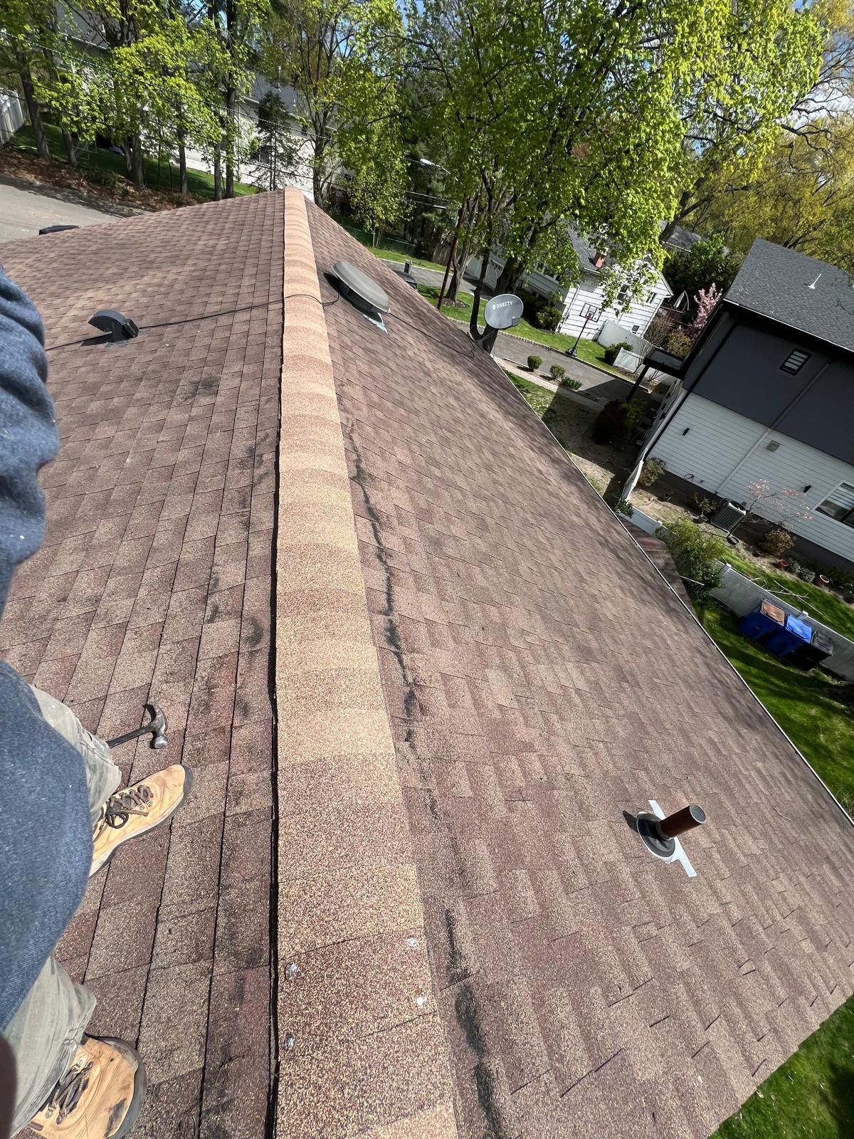Person on brown shingle roof with a ridge cap, trees, and houses in the background.
