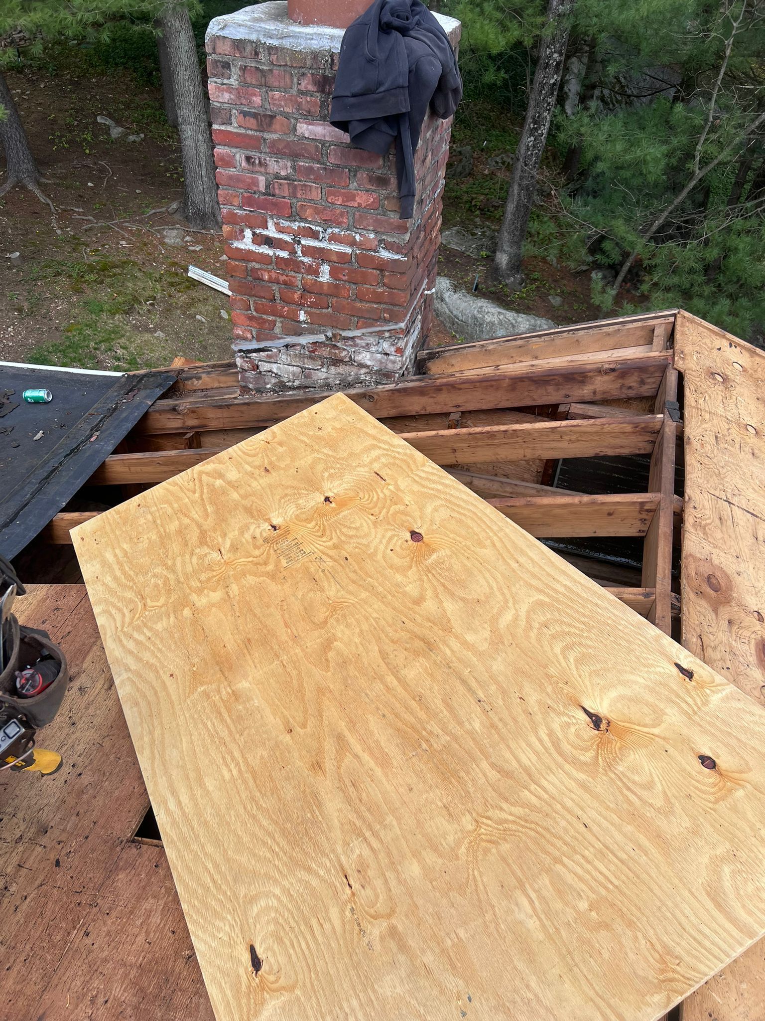Rooftop with a damaged wooden frame and a brick chimney. A sheet of plywood rests on the frame.
