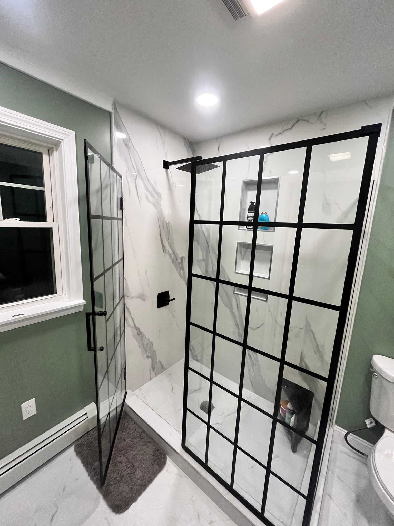 Modern bathroom with black-framed glass shower. Marble-look tile, green walls, and a dog inside the shower.