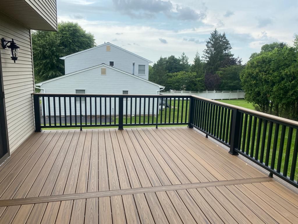 Wooden deck with black railing overlooking a grassy yard with a house in the background on a cloudy day.
