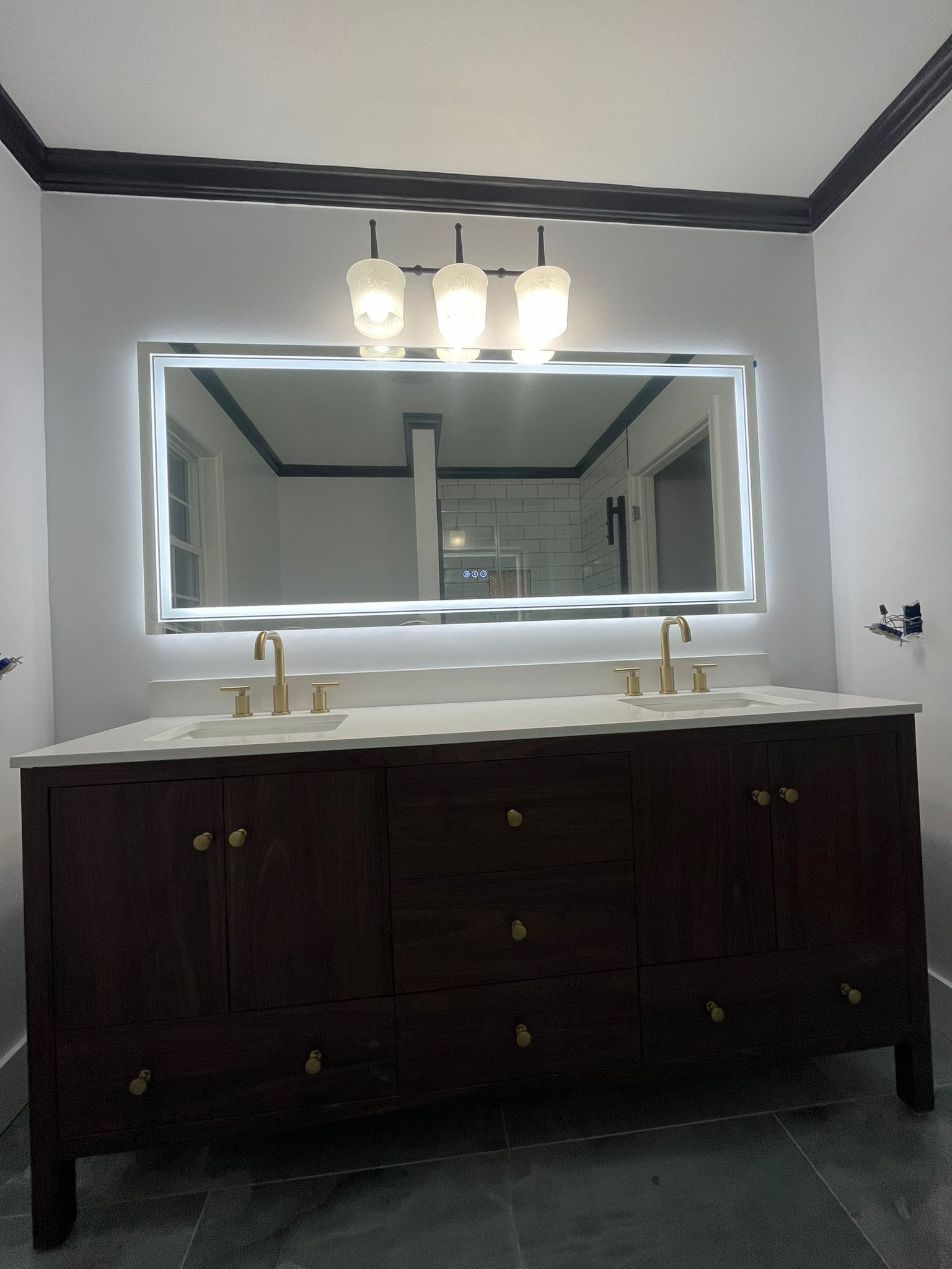 Bathroom with a dark brown vanity, large mirror with LED lights, gold fixtures, and a three-bulb light fixture.