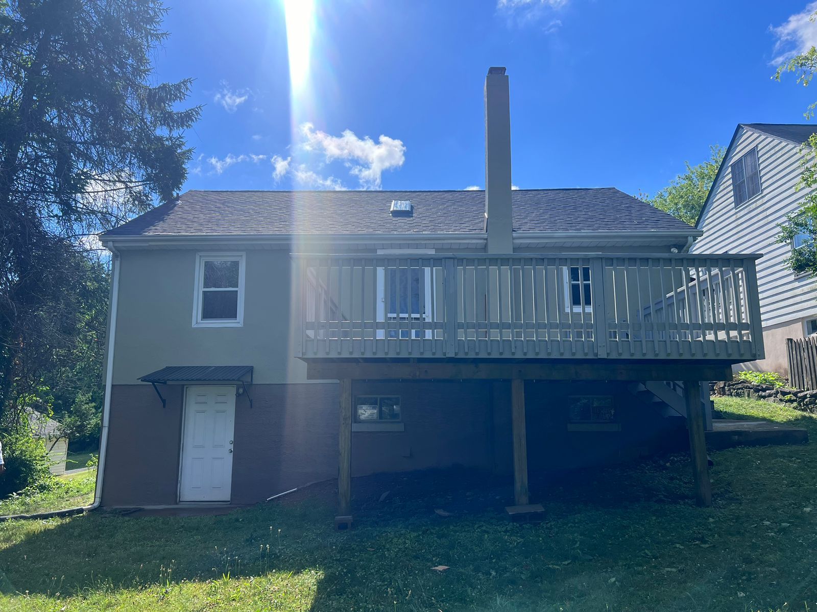 Rear view of a two-story house with a wooden deck and chimney on a sunny day.