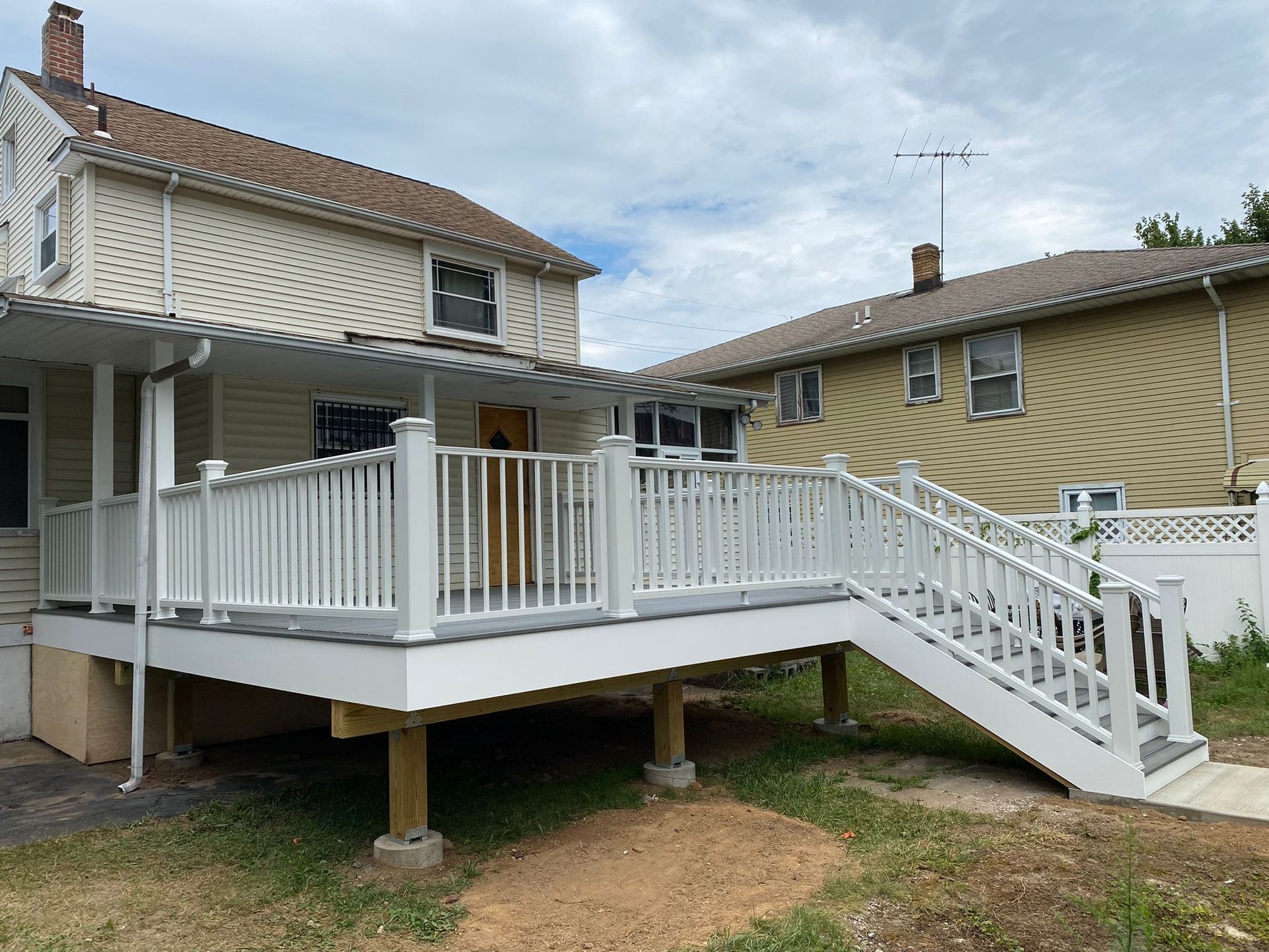 White deck with stairs, attached to a beige house with a covered porch, set outdoors on a cloudy day.