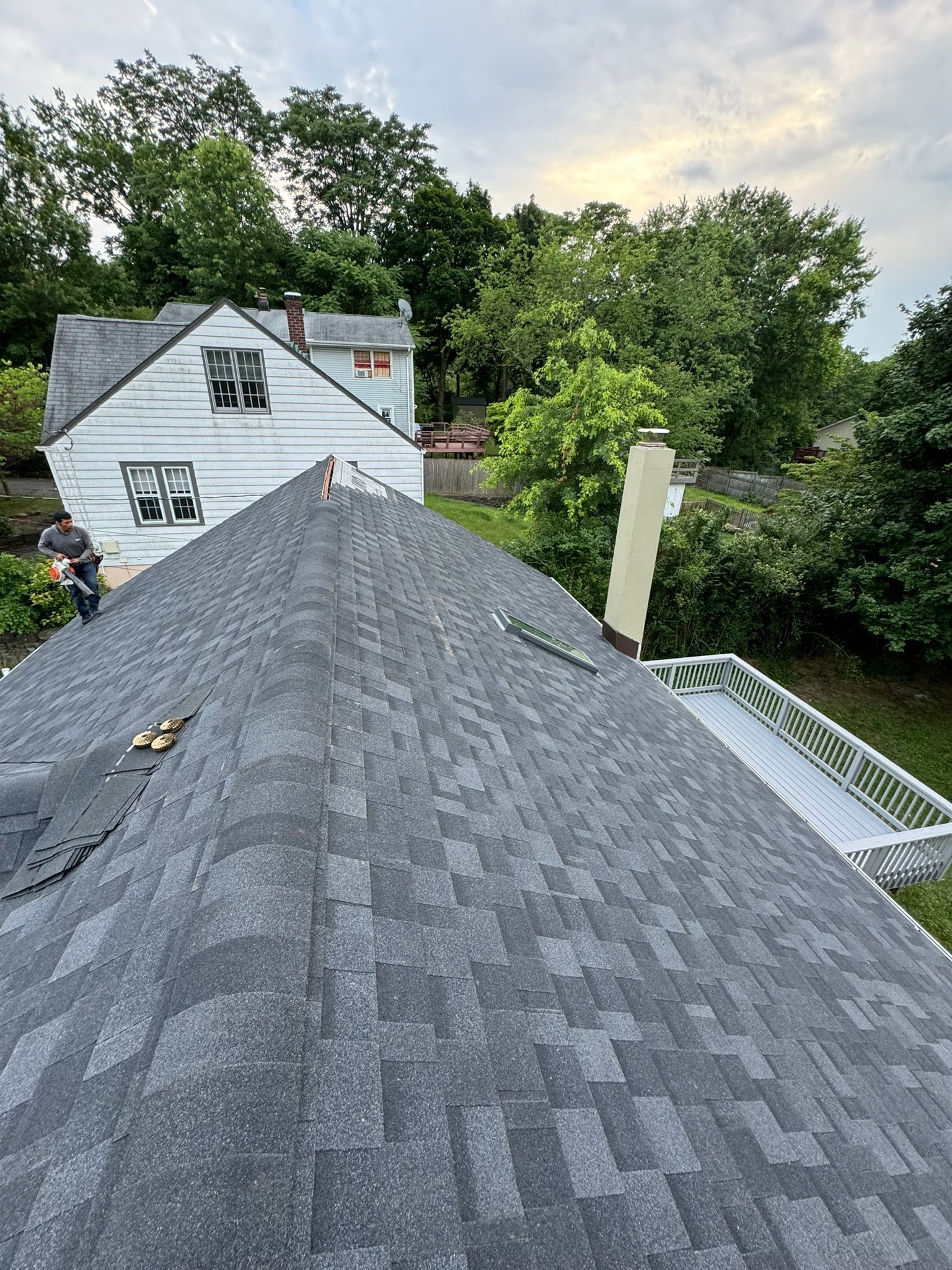 Gray shingled roof, workers, chimney, and two houses in the background, all under a cloudy sky.