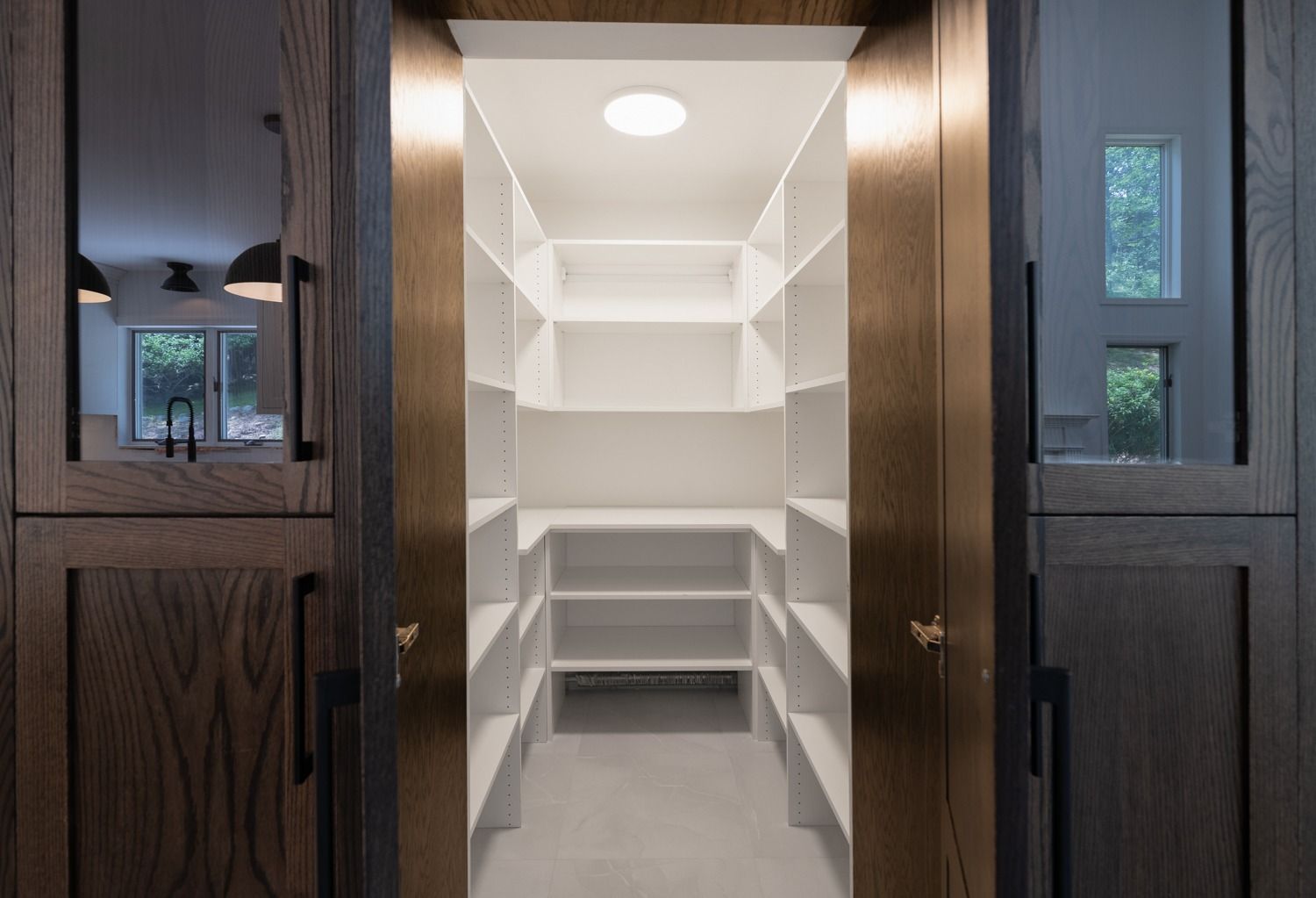 Walk-in pantry with white shelves and lighting, flanked by dark wooden cabinets with mirrors and windows.