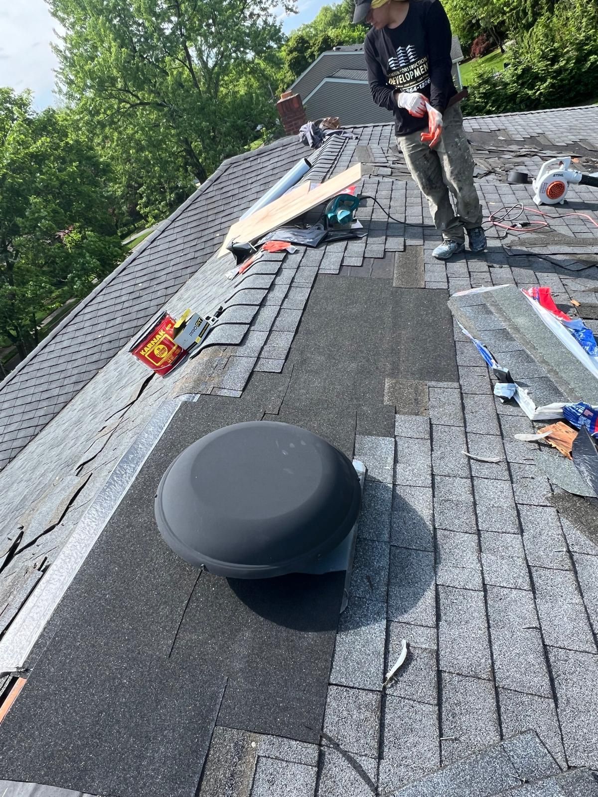 Man repairing roof; shingles removed near a large, round vent. Daylight.