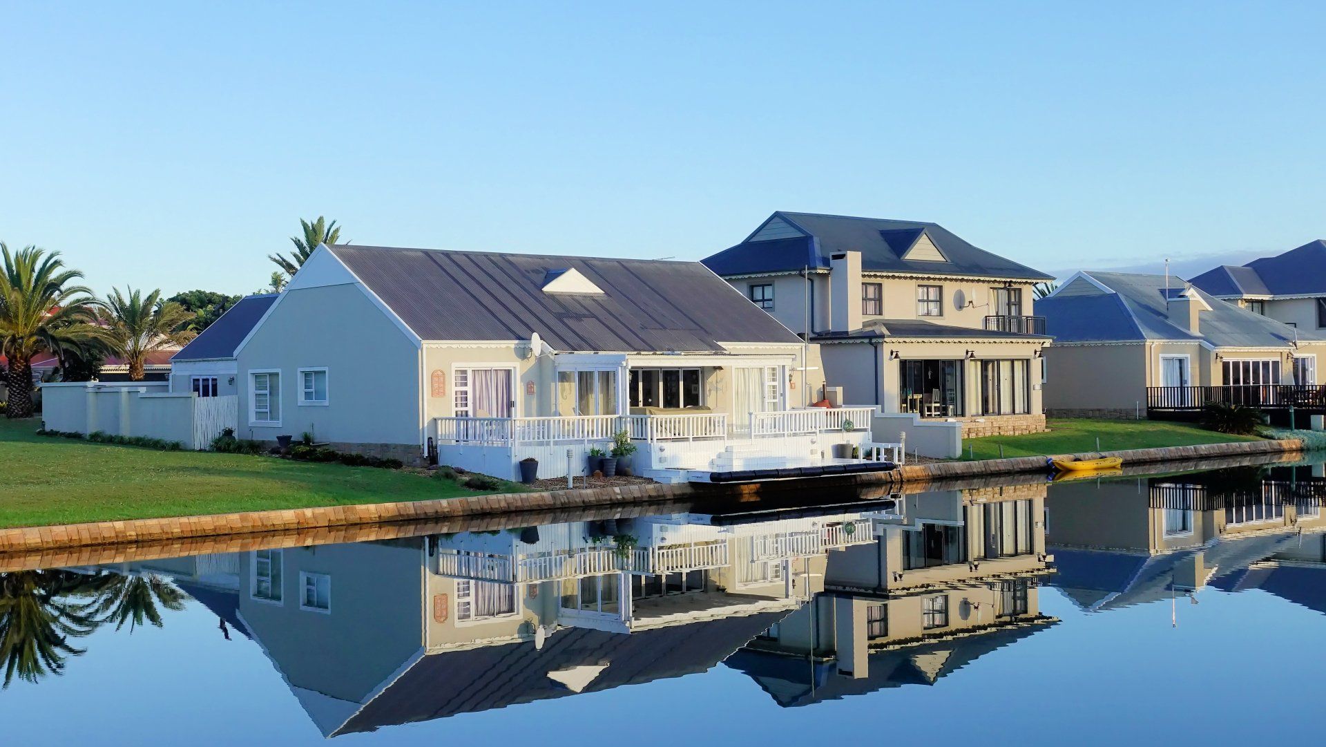 Houses on a canal with reflections in the water, blue sky, green lawn.