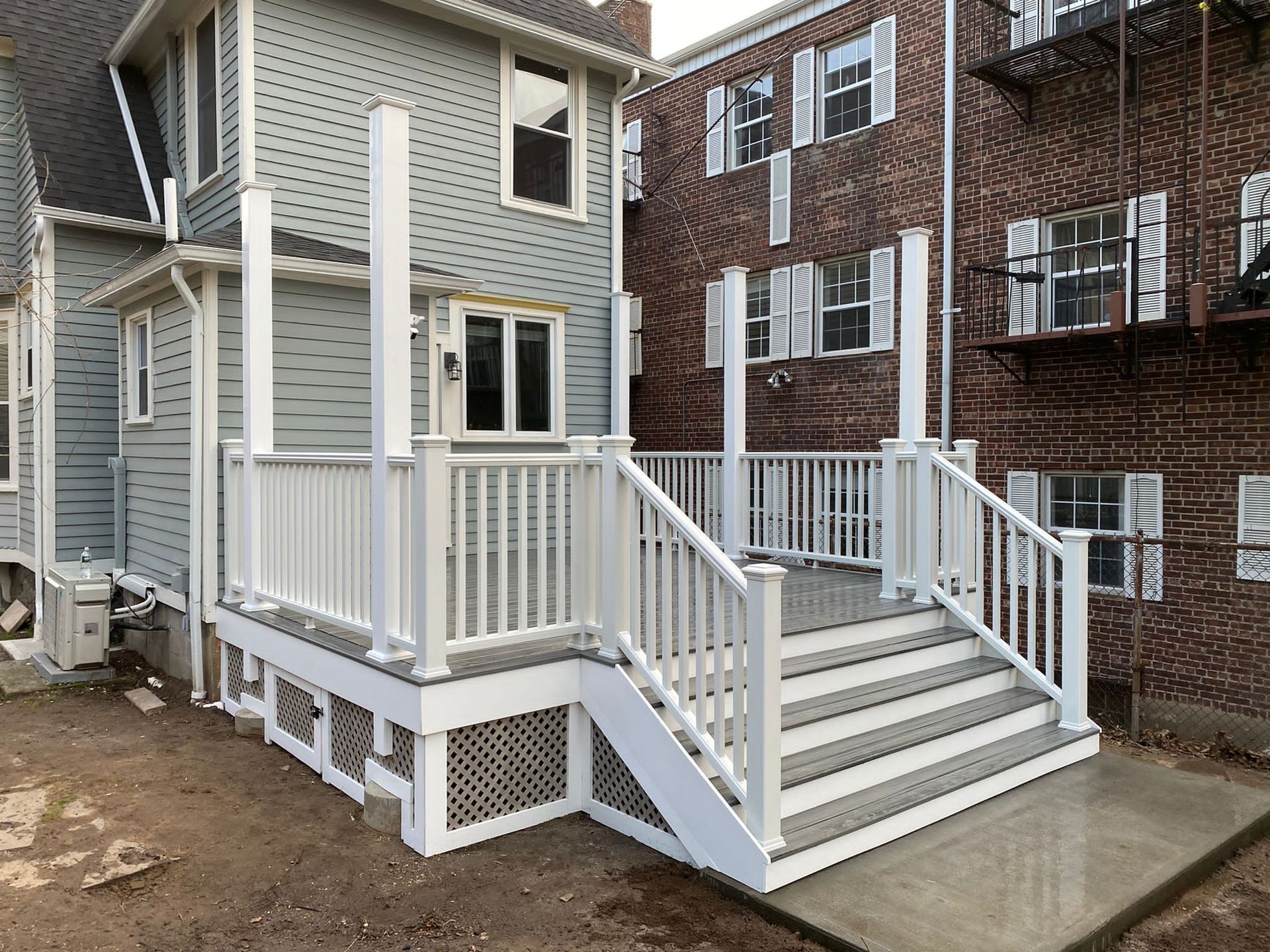 White deck with stairs, railings, and posts against a gray-sided building, next to a brick building.