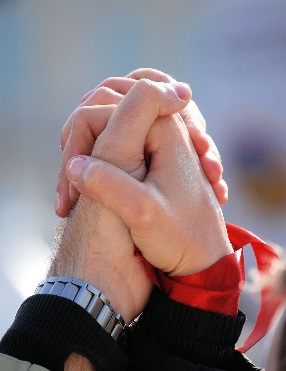 Two people hold hands in a firm grip, one wearing a watch, with a hint of a red garment visible in the background.