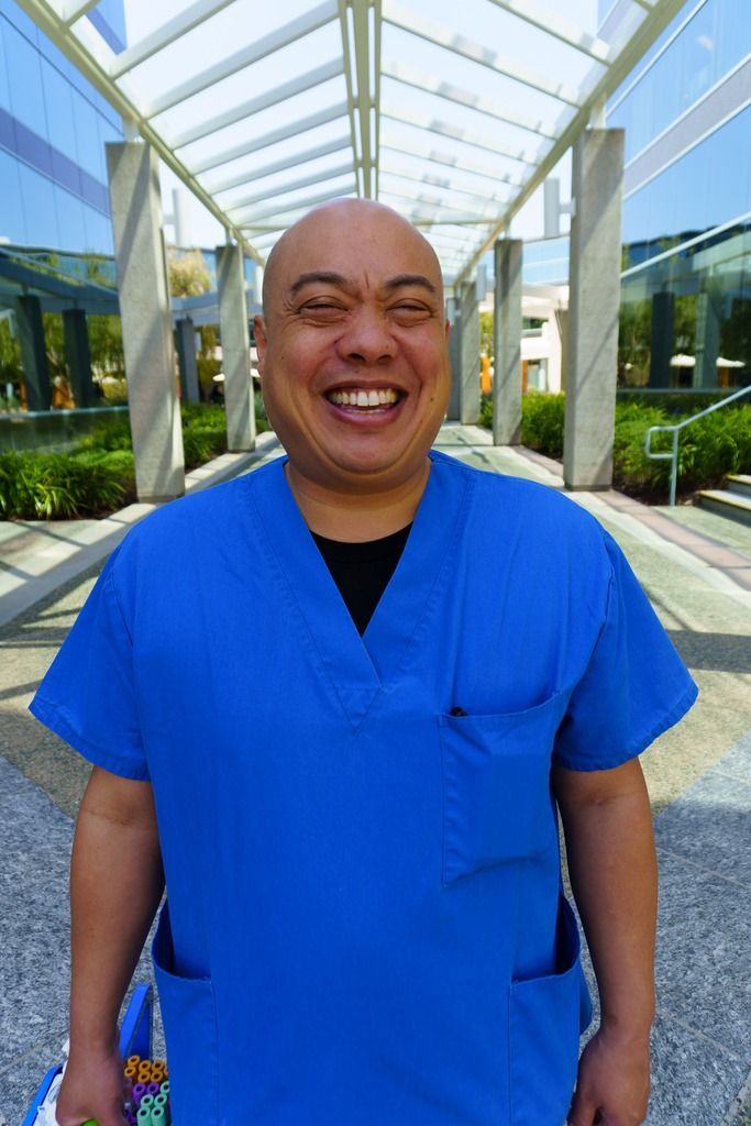 A smiling individual wearing bright blue scrubs stands outdoors under a modern, glass-covered walkway.