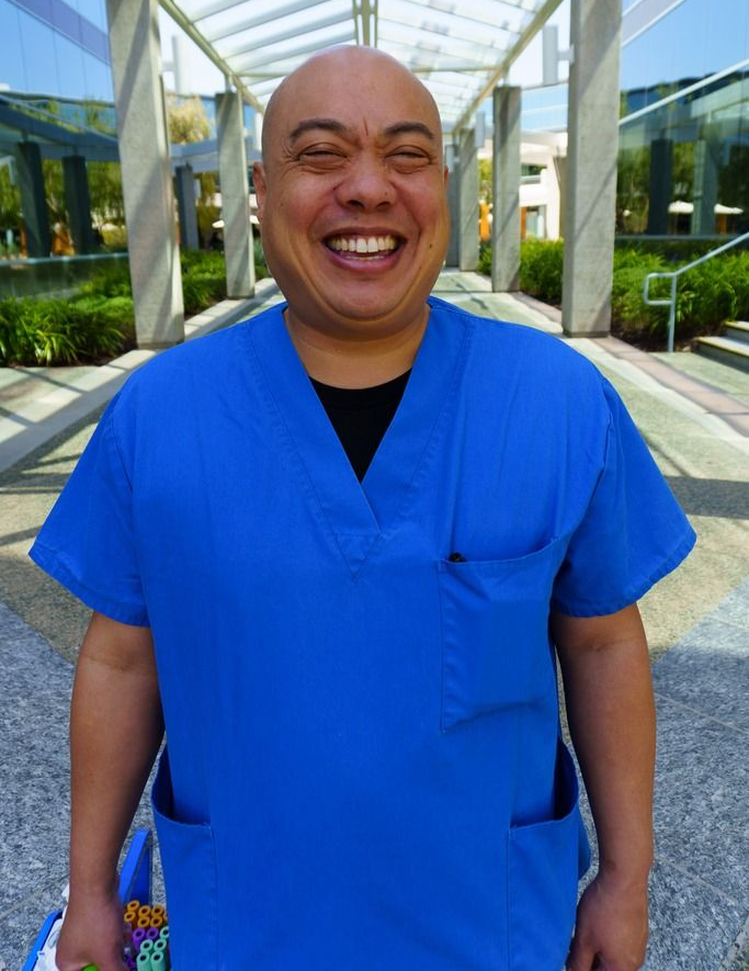 A smiling individual wearing bright blue scrubs stands outdoors under a modern, glass-covered walkway.
