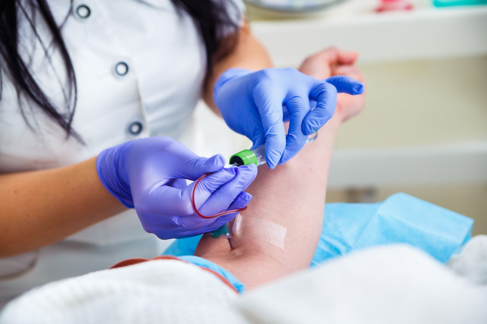 A medical professional wearing blue gloves performs a blood draw on a patient's arm.