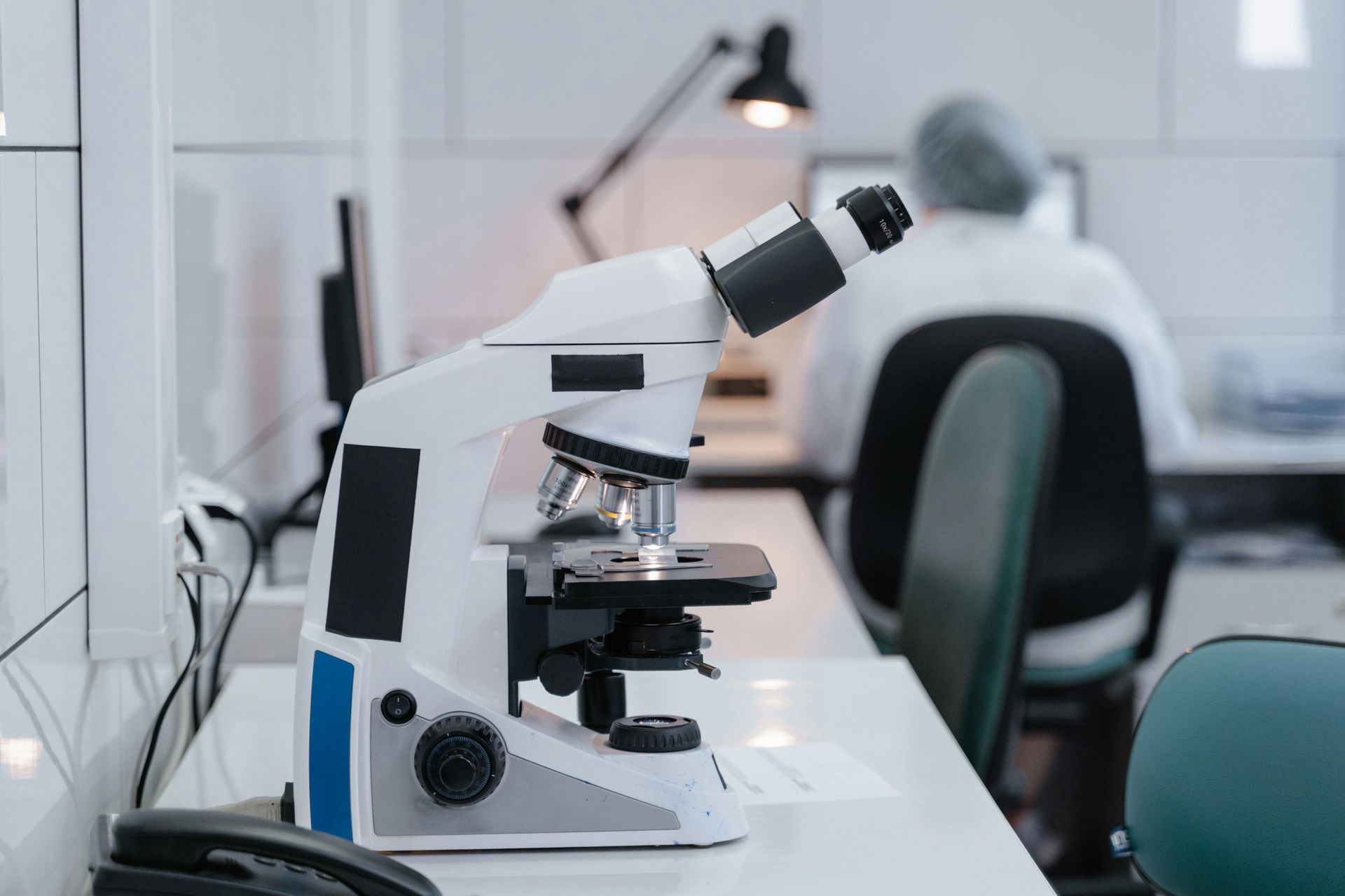 A laboratory microscope sits on a white desk in the foreground, with a person working at a computer in