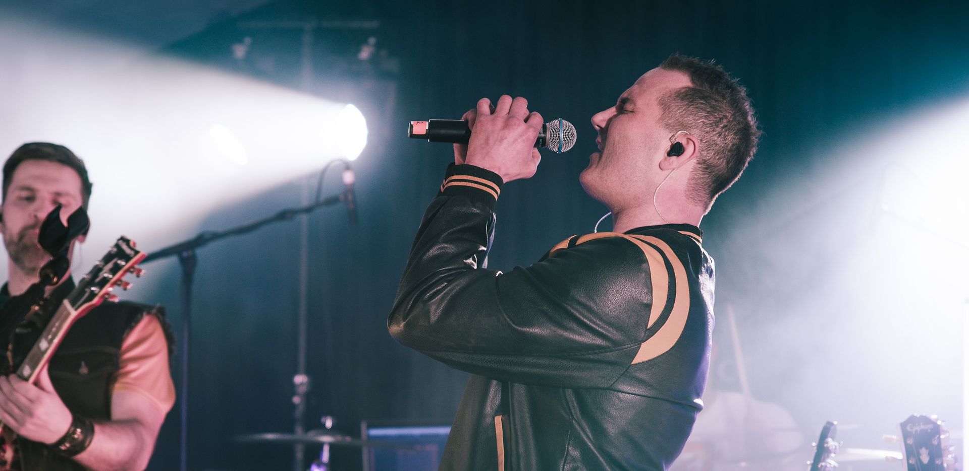 En mann synger i en mikrofon på en scene mens en annen mann spiller gitar.