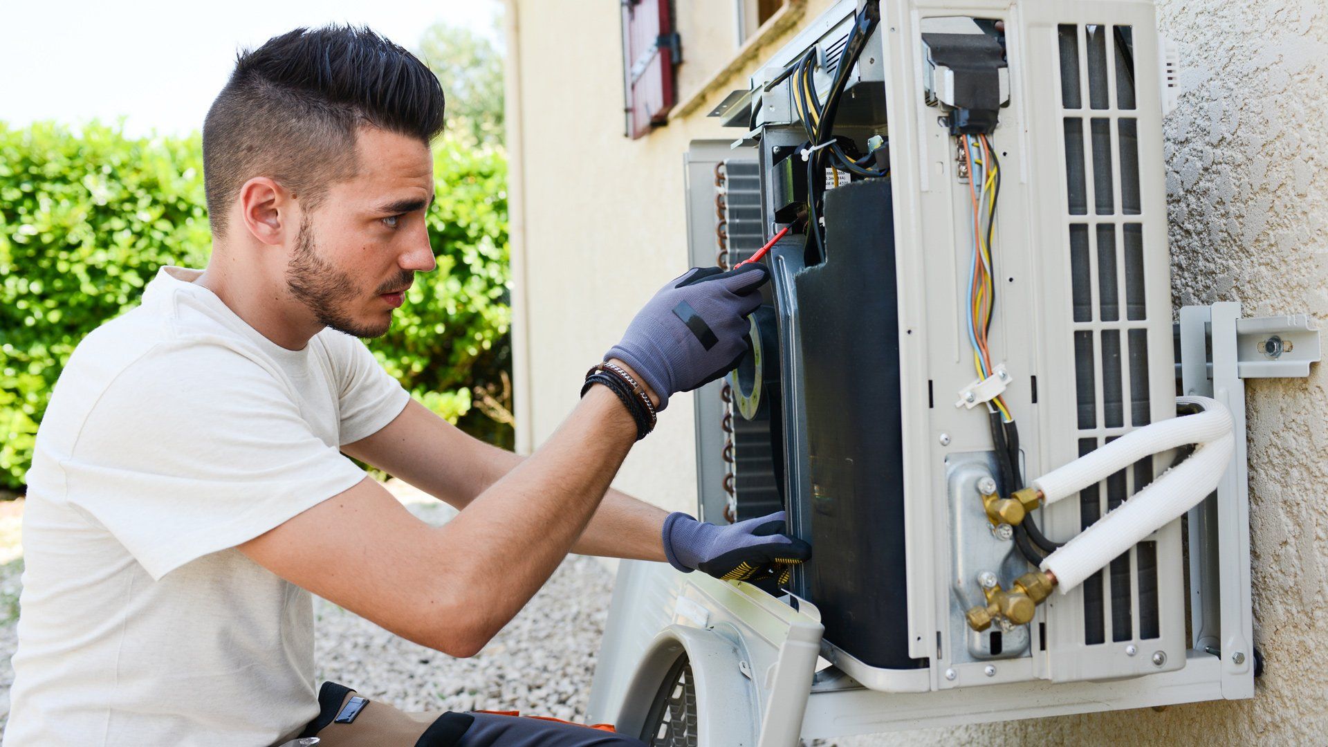 Technician inspecting an outdoor air conditioner unit with tools outside a house