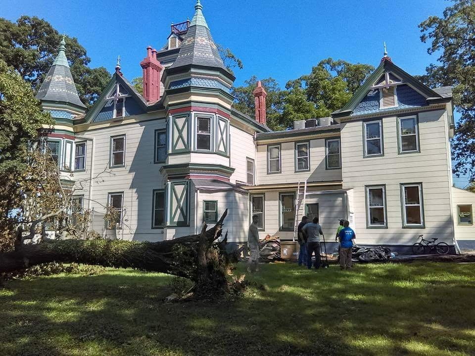 Victorian house with turret and porch on a sunny day, two people standing on the lawn.