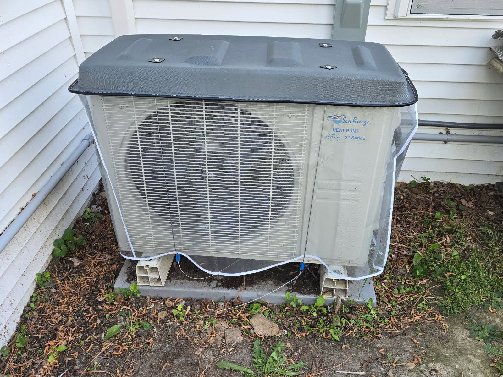 Outdoor HVAC condenser unit beside a house, with a protective wire cage and dark cover on top