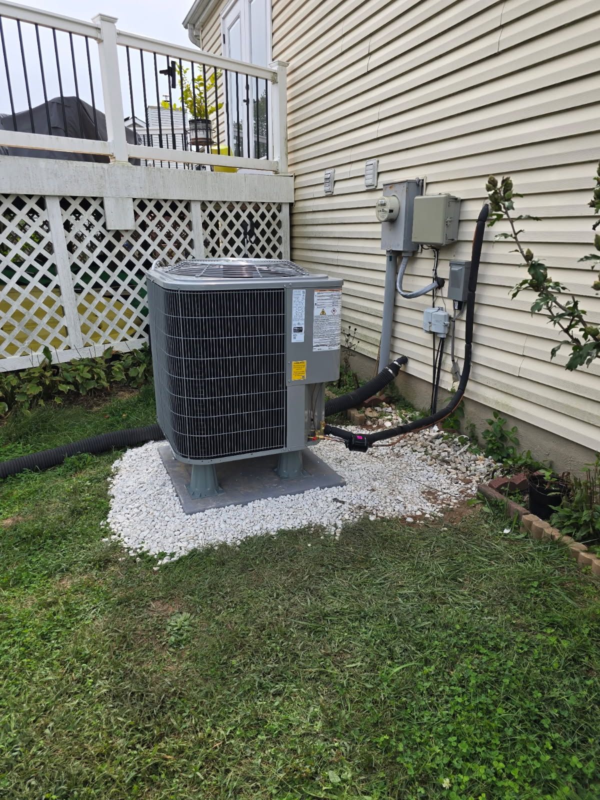 Outdoor air conditioner unit beside beige house wall on gravel pad, with grass and lattice fence nearby