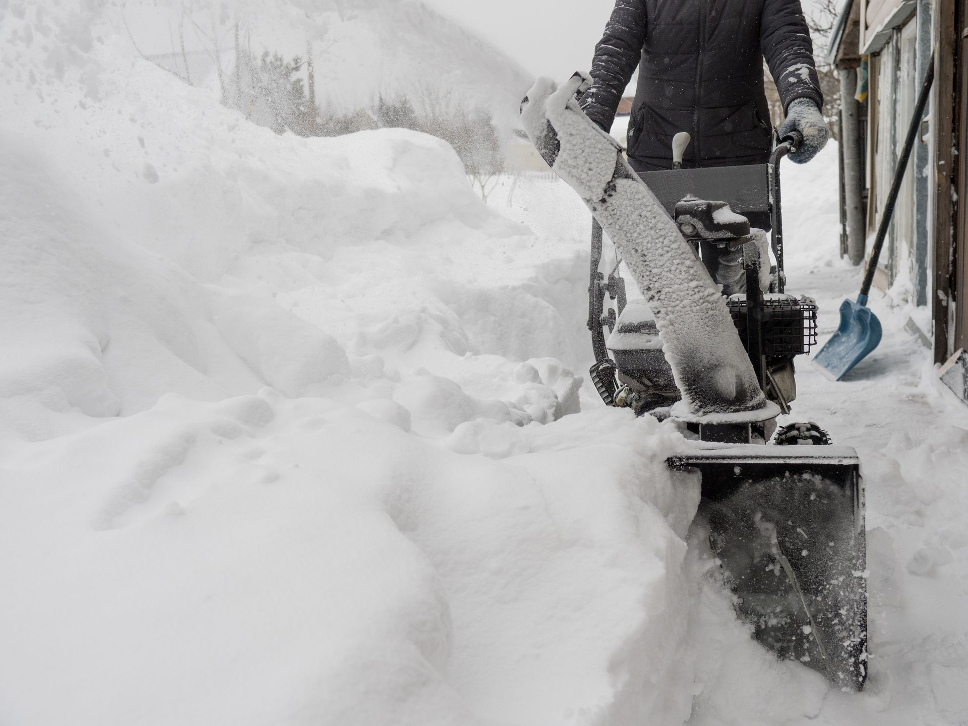 Person using a snowblower to clear deep snow near a building in winter.