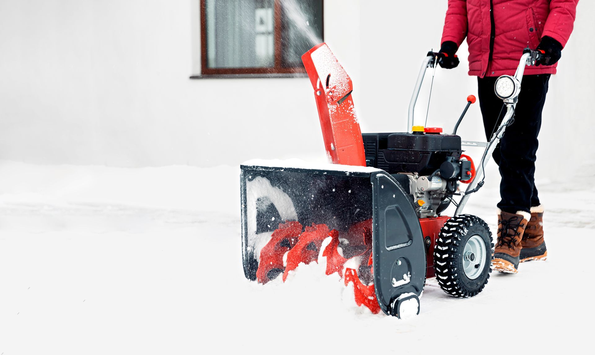 Person in red jacket using a red and black snowblower to clear snow in front of a building.