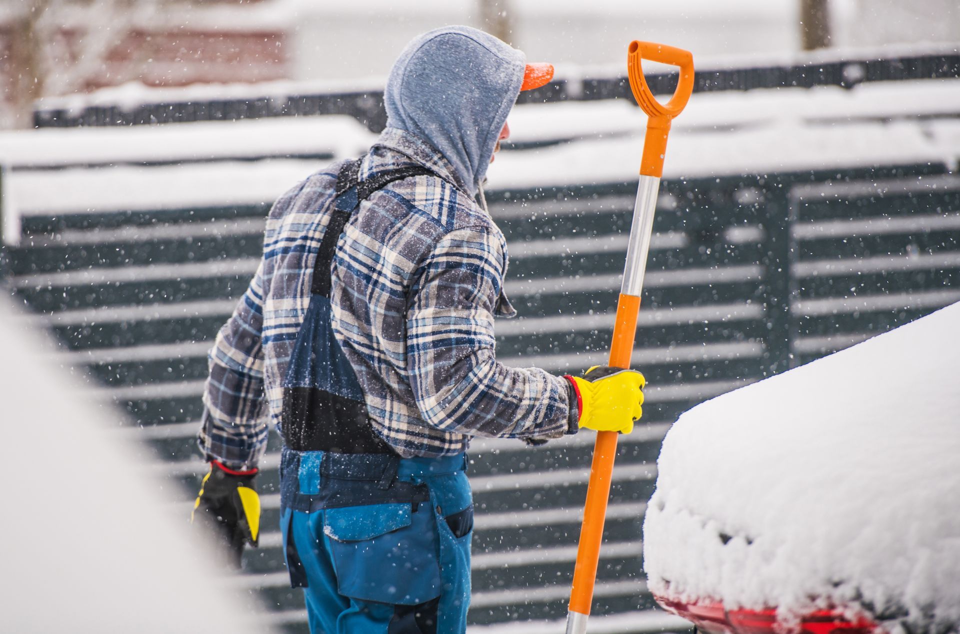 Person shoveling snow, wearing blue overalls, plaid shirt, hooded sweatshirt, and yellow gloves; snow falling.