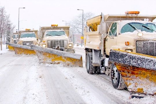 Snowplows clearing snow from a road; winter scene, yellow blades, trucks in a row.