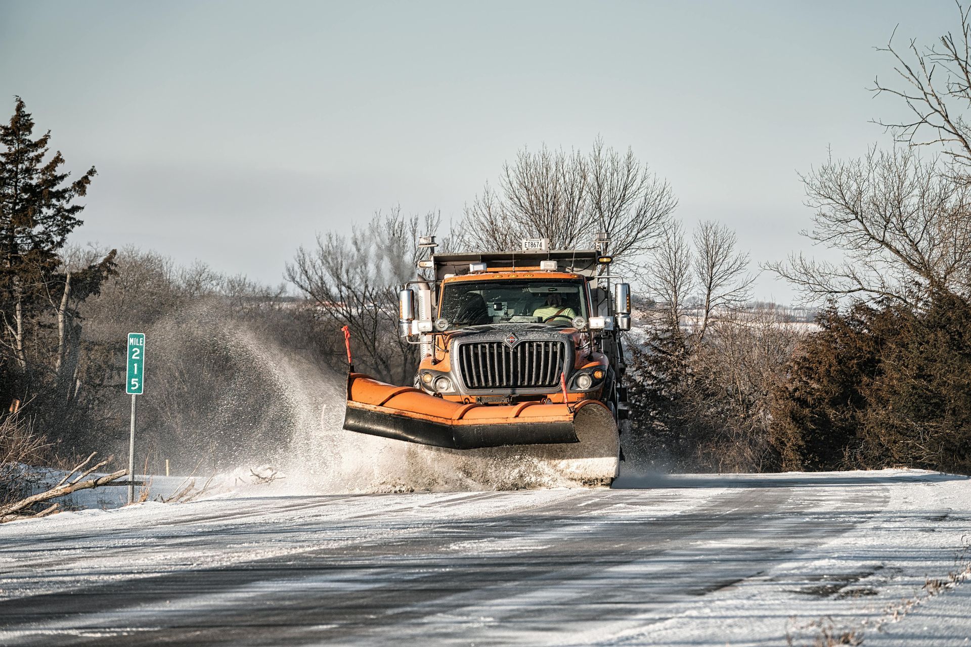 Snowplow on a snowy road, orange blade, spraying slush, trees in background.